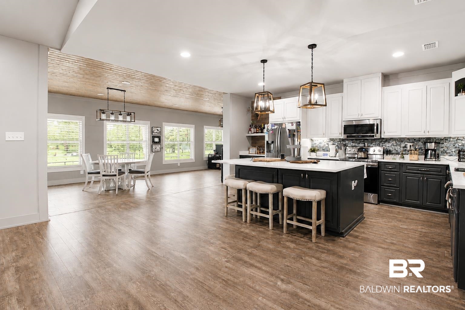 9825 Wolf Creek Ridge Elberta, AL 36530 - Photo 33 of 76 Open-concept kitchen and dining area featuring wood-finish flooring, a large kitchen island with seating, a patterned backsplash, white upper cabinetry, and dark lower cabinetry