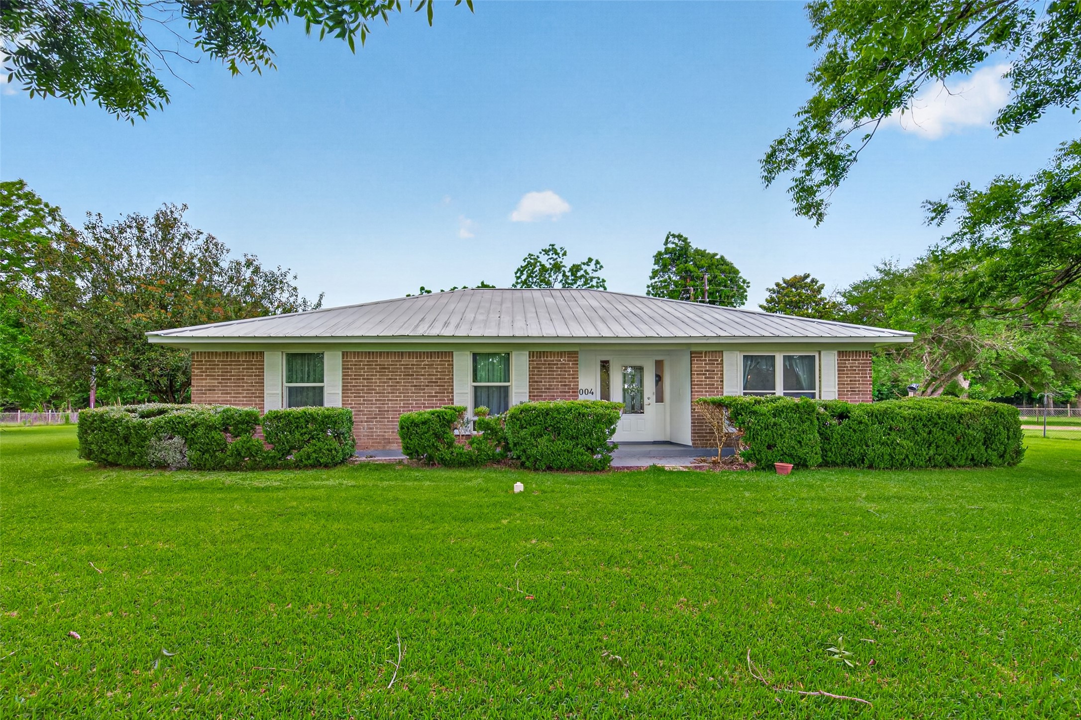 This charming one-story brick home welcomes you with its classic exterior, a striking metal roof, and lush, manicured landscaping that lines the front facade. A generous, well-kept lawn stretches across the property, framed by mature trees that add shade and natural beauty. The covered front entry offers a warm and inviting first impression.