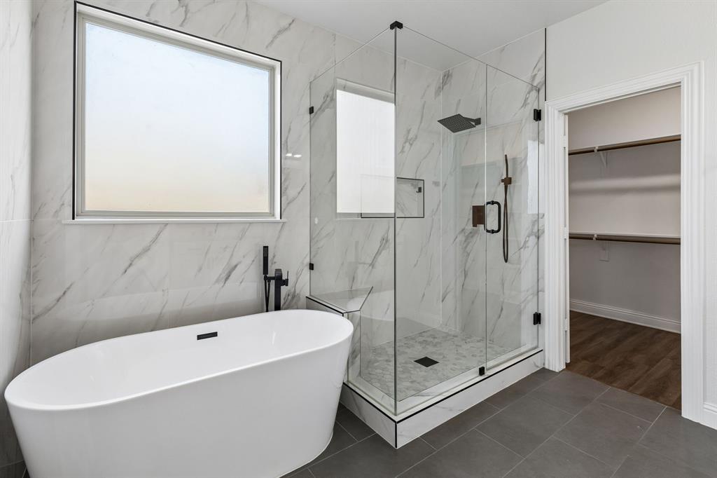 537 Hunt Farms Road Fate, TX 75189 - Photo 21 of 36 Freestanding soaking tub with a floor-mounted faucet, glass-enclosed shower with a built-in bench, wall-to-wall marble-patterned tile, and gray floor tile