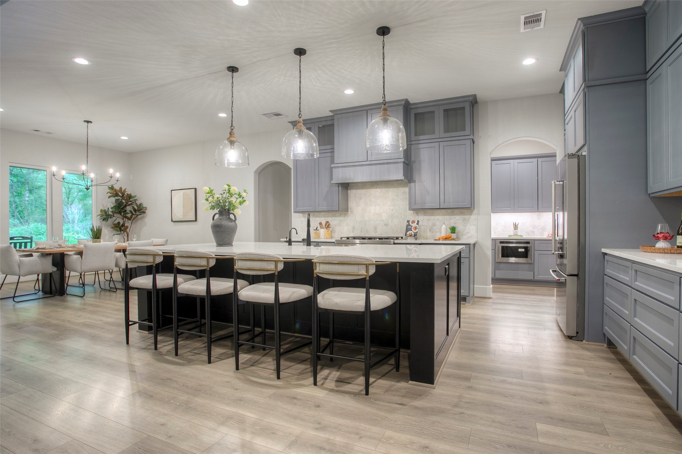 12915 Oak Barrel Road Willis, TX 77378 - Photo 12 of 46 This spacious, modern kitchen features sleek gray cabinetry, a large island with seating, stainless steel appliances, and elegant pendant lighting. The open layout flows into a bright dining area with large windows overlooking greenery, creating a perfect space for entertaining.
