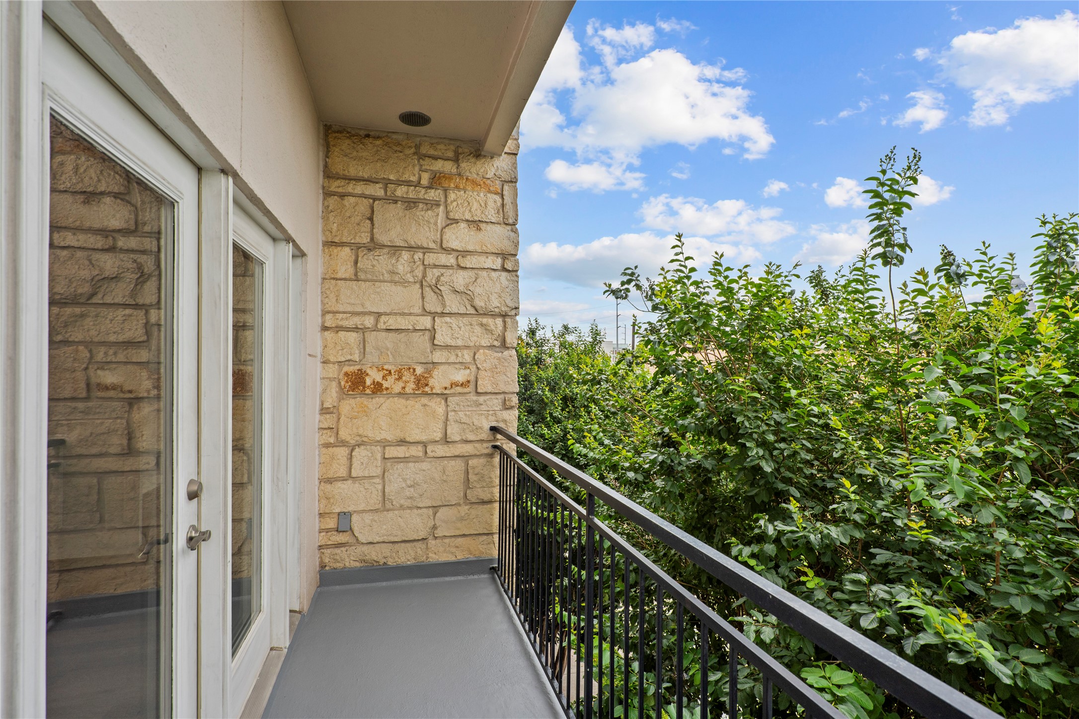 1900 Genesee Street, Unit 203 Houston, TX 77006 - Photo 22 of 30 This photo shows a cozy balcony with a stone wall, French doors, and a black metal railing, overlooking lush greenery under a bright sky.