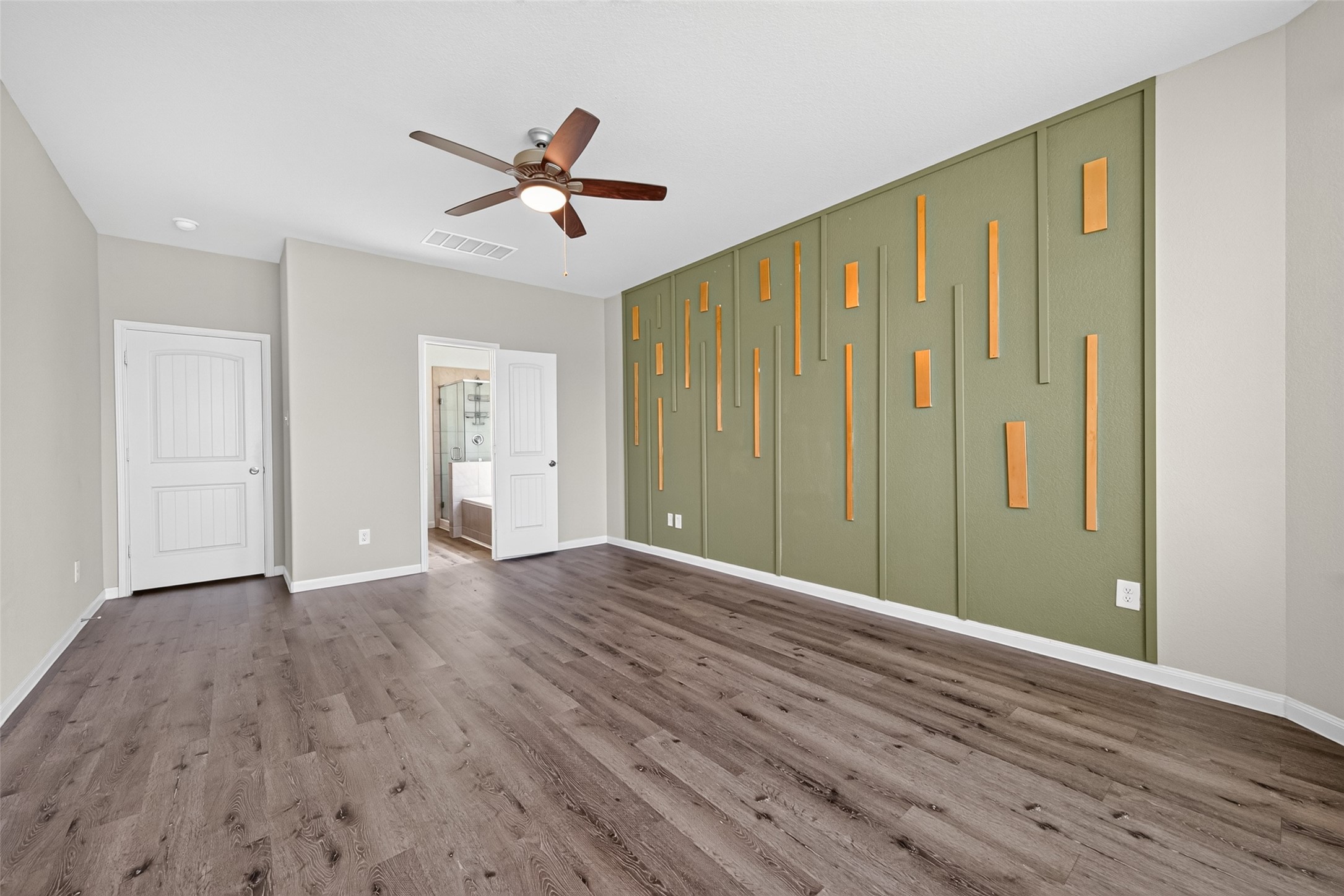 2526 Sturgeon Way Conroe, TX 77384 - Photo 21 of 49 This spacious bedroom features a modern accent wall with vertical wooden elements on a green background, wood flooring, and a ceiling fan. It includes a doorway to a bathroom and a closet, offering a stylish and functional living space.