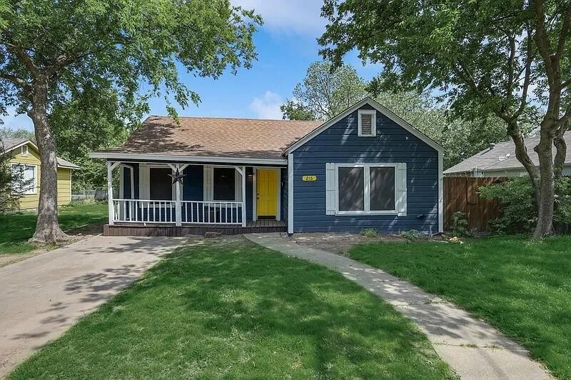 216 East Division Street Pilot Point, TX 76258 - Photo 1 of 17 Blue siding exterior featuring a covered front porch with white railing, a vibrant yellow front door, and a brown shingle roof