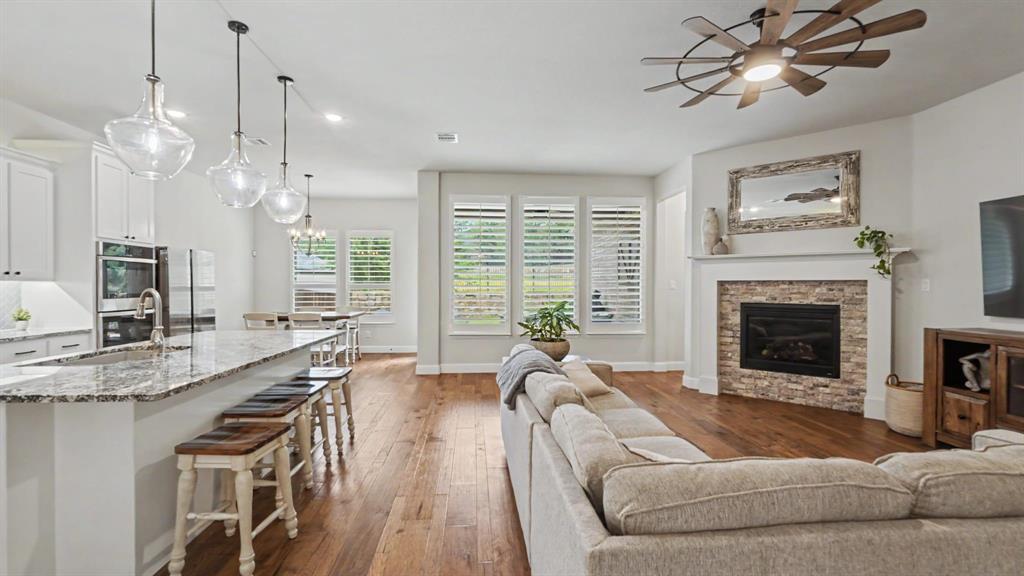 5213 Ravine Ridge Court Flower Mound, TX 76262 - Photo 1 of 40 Open-concept living space featuring hardwood floors, a stone-surround fireplace with a white mantel, and a kitchen island with pendant lighting. Windows overlook backyard from the dining area, living, and kitchen.