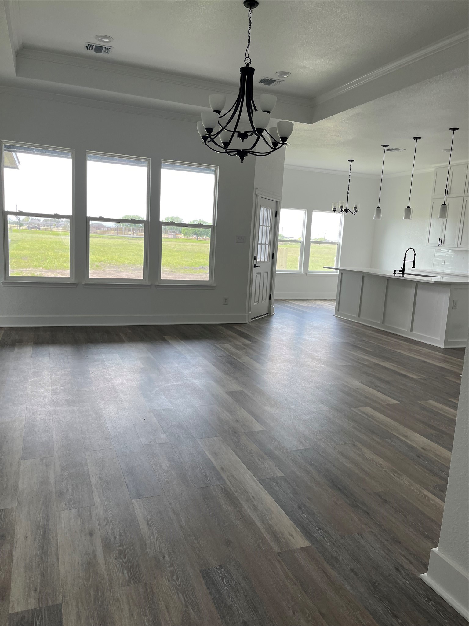 33416 FM 521 Road Angleton, TX 77515 - Photo 7 of 21 This photo showcases a bright, open-concept living space with large windows, medium gray/wood water proof plank flooring, and modern light fixtures. It features a spacious kitchen area with a white island and pendant lighting, offering a view of a green outdoor space.