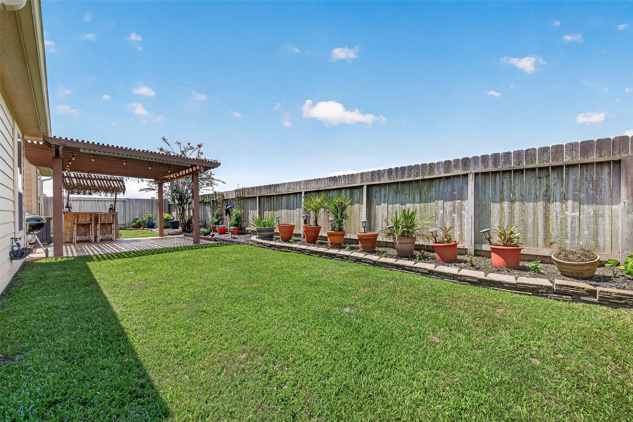 4910 Culver Lane Rosenberg, TX 77471 - Photo 47 of 50 This backyard features a spacious grassy area with a charming pergola perfect for outdoor dining. A row of potted plants lines the wooden fence, creating a colorful and inviting atmosphere.