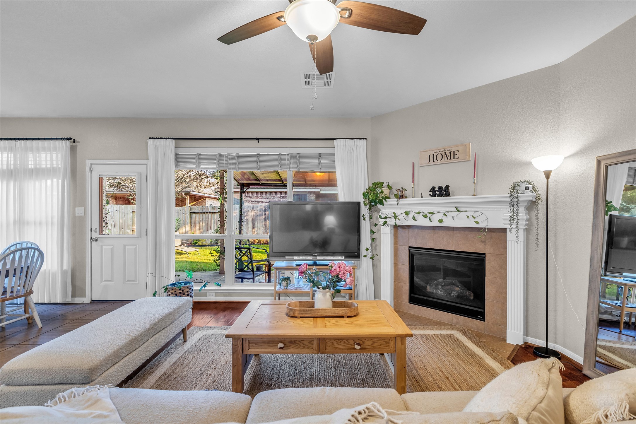 155 Magnolia Grove Lane Conroe, TX 77384 - Photo 4 of 25 Cozy living room with a neutral color palette, featuring a fireplace, ceiling fan, and large windows allowing natural light. The space includes a comfortable seating area, a wall-mounted TV, and a view of the backyard patio through glass doors. Ideal for relaxation and entertaining.