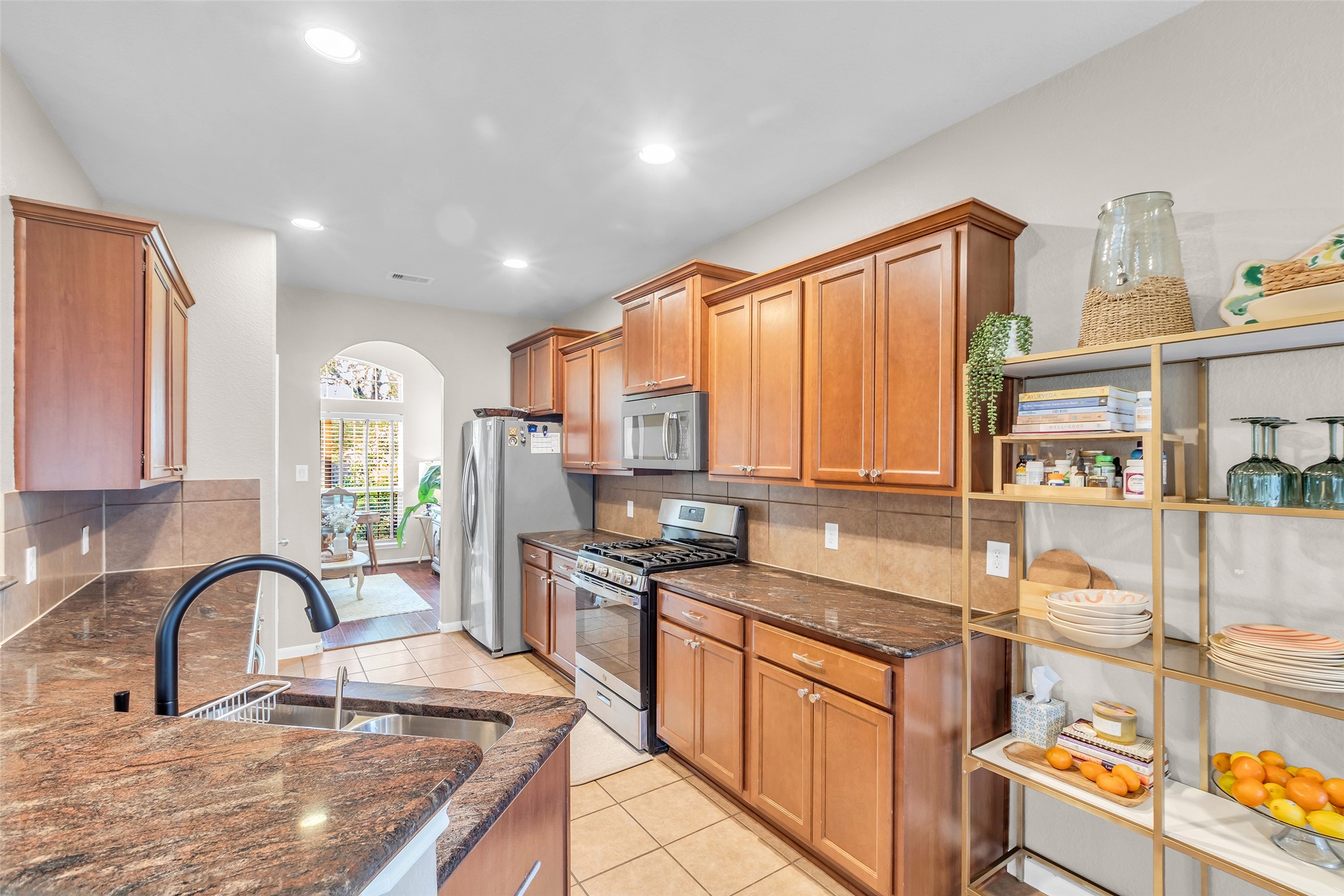 155 Magnolia Grove Lane Conroe, TX 77384 - Photo 7 of 25 This kitchen features warm wood cabinetry, granite countertops, and stainless steel appliances. It includes a modern sink, open shelving for storage, and an arched doorway leading to a sitting area with large windows, filling the space with natural light.