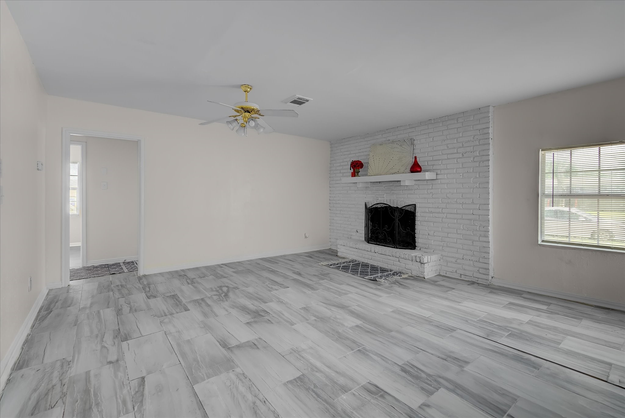 418 Azalea Street Lake Jackson, TX 77566 - Photo 11 of 34 This room features light gray flooring and a white brick fireplace with a mantel. It includes a ceiling fan, a large window for natural light, and a doorway leading to another space, offering a clean and versatile living area.
