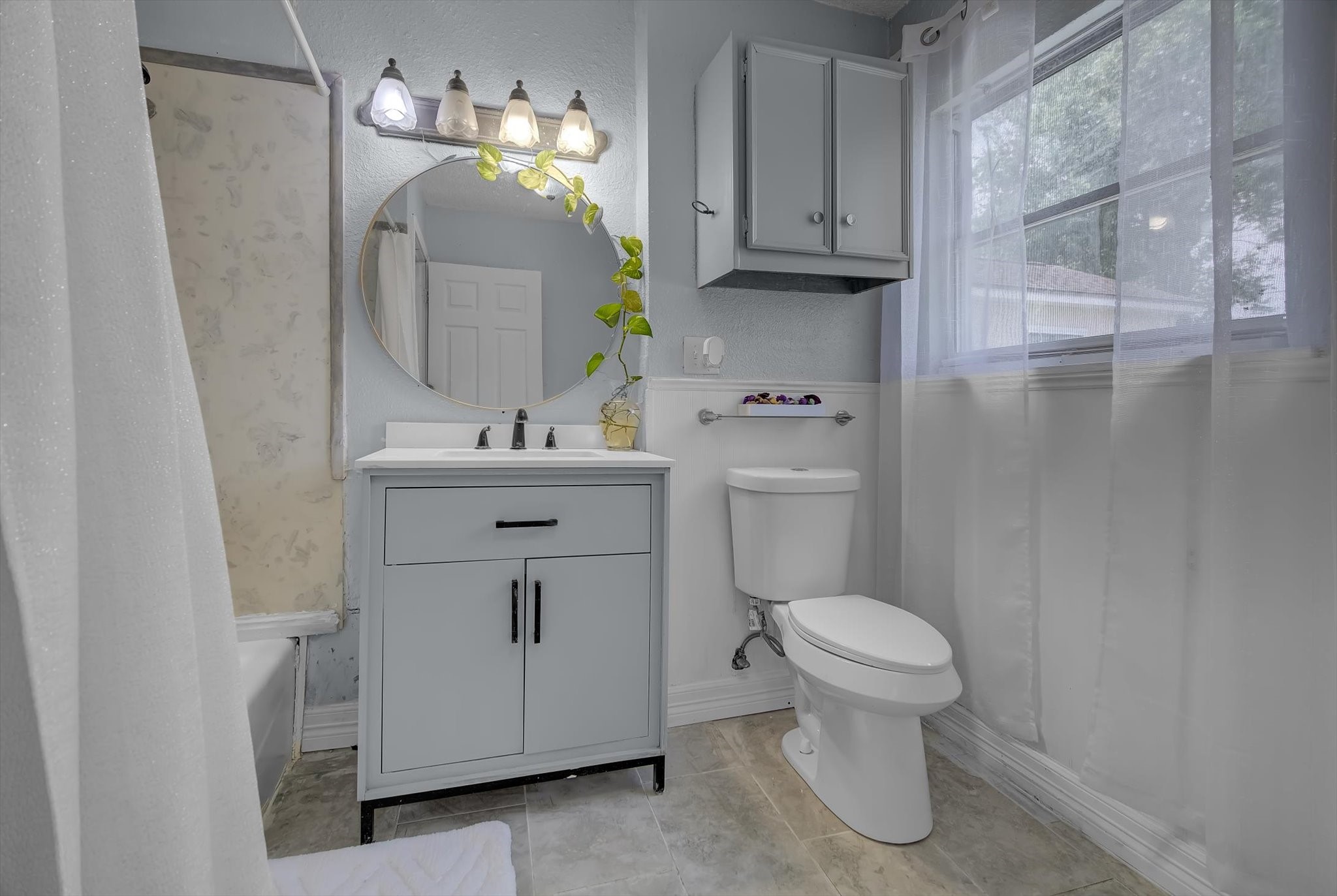 418 Azalea Street Lake Jackson, TX 77566 - Photo 5 of 34 This bathroom features a modern design with a light gray vanity, round mirror, and overhead lighting. It includes a shower-tub combo with a curtain, a toilet, and a window with sheer curtains for natural light. The walls are a soft gray, complementing the clean and fresh look.