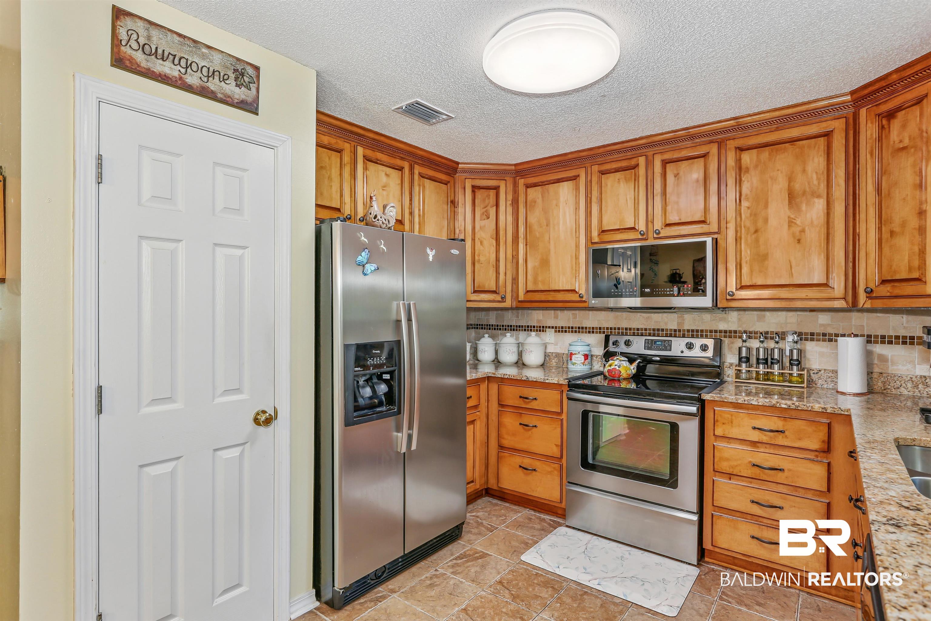 32334 Riverlake Road Seminole, AL 36574 - Photo 22 of 47 Kitchen featuring appliances with stainless steel finishes, light stone counters, brown cabinetry, a textured ceiling, and backsplash
