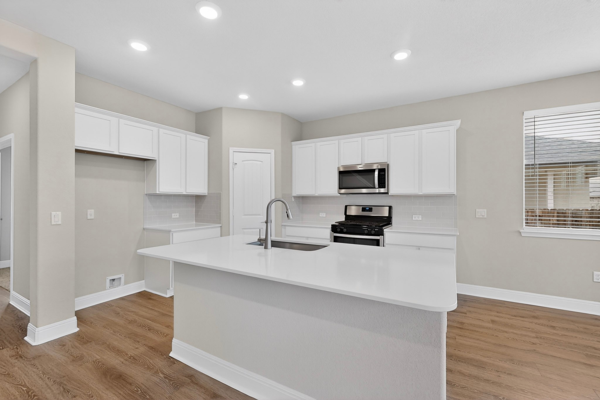 16809 Guido Cove Pflugerville, TX 78660 - Photo 11 of 40 Open-concept kitchen featuring an island with an undermount sink and gooseneck faucet, white shaker cabinetry, stainless steel appliances including a microwave and range, and wood-finish flooring