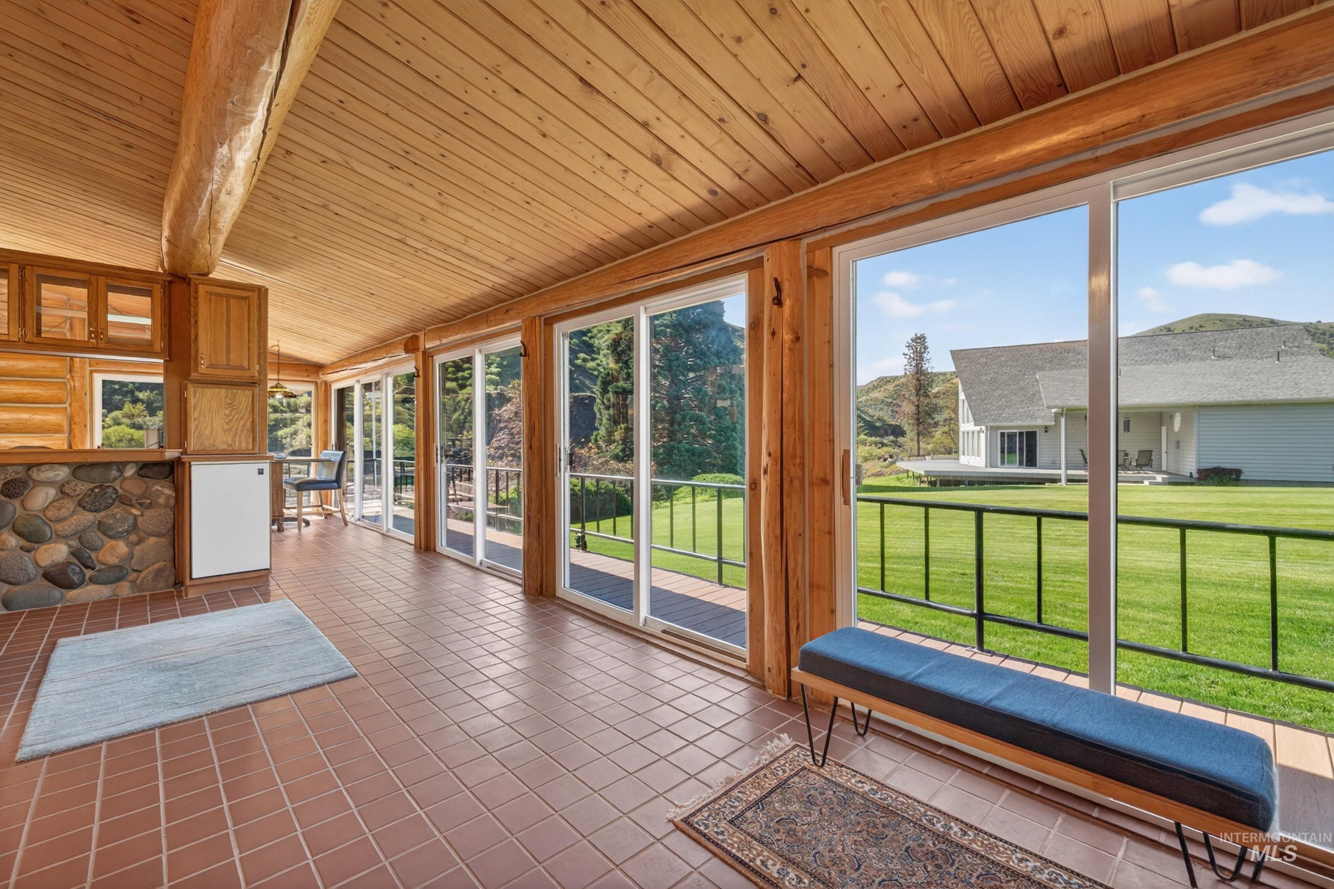 215 Doumecq Road White Bird, ID 83554 - Photo 16 of 50 Sunroom featuring extensive natural wood paneling, exposed wood beams, brown tile flooring, a built-in stone bar, and multiple sliding glass doors opening to a deck