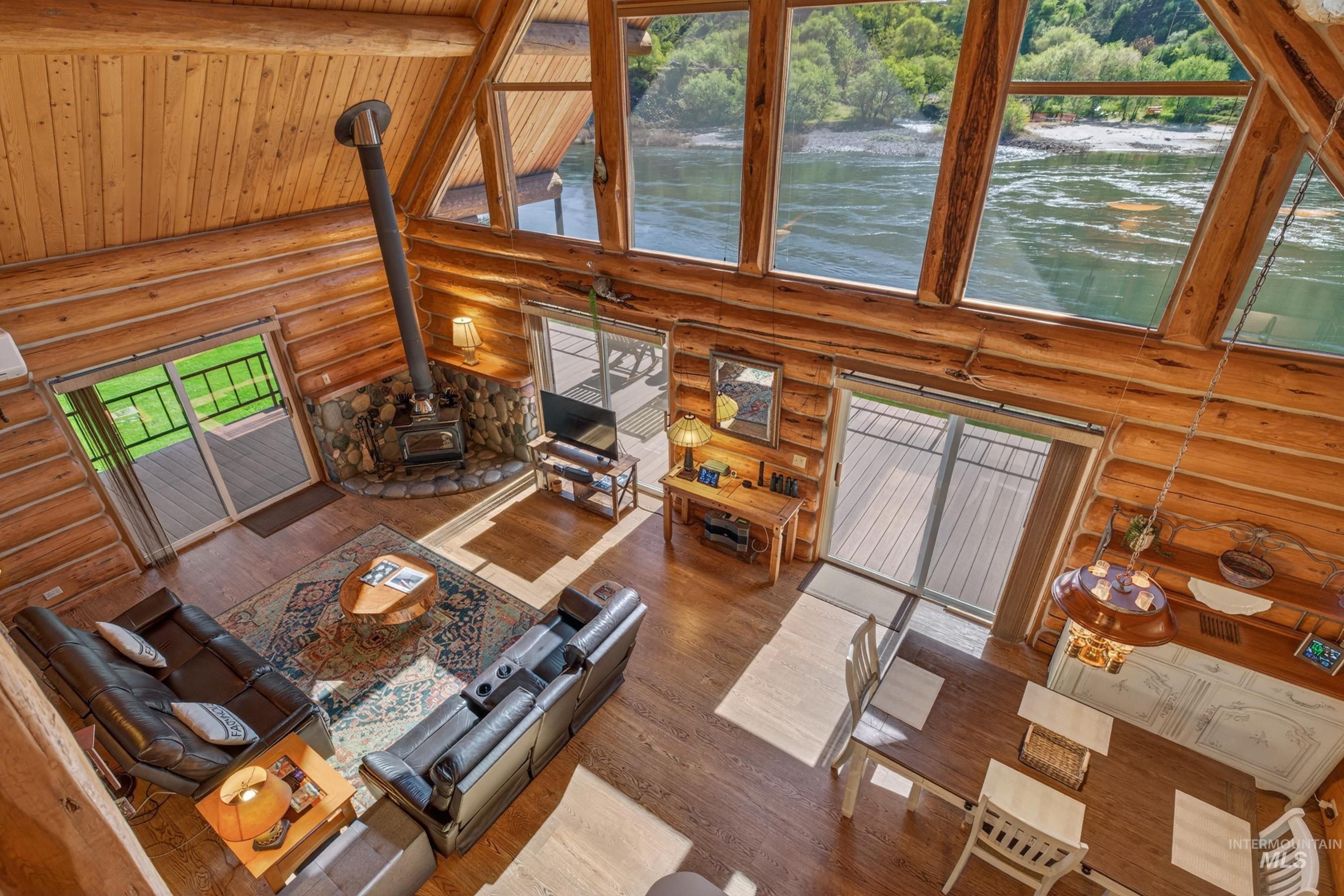 215 Doumecq Road White Bird, ID 83554 - Photo 6 of 50 Expansive open-concept room featuring a wood-burning stove with a stone hearth, vaulted ceilings with exposed wood beams, and a wall of windows showcasing a river backdrop