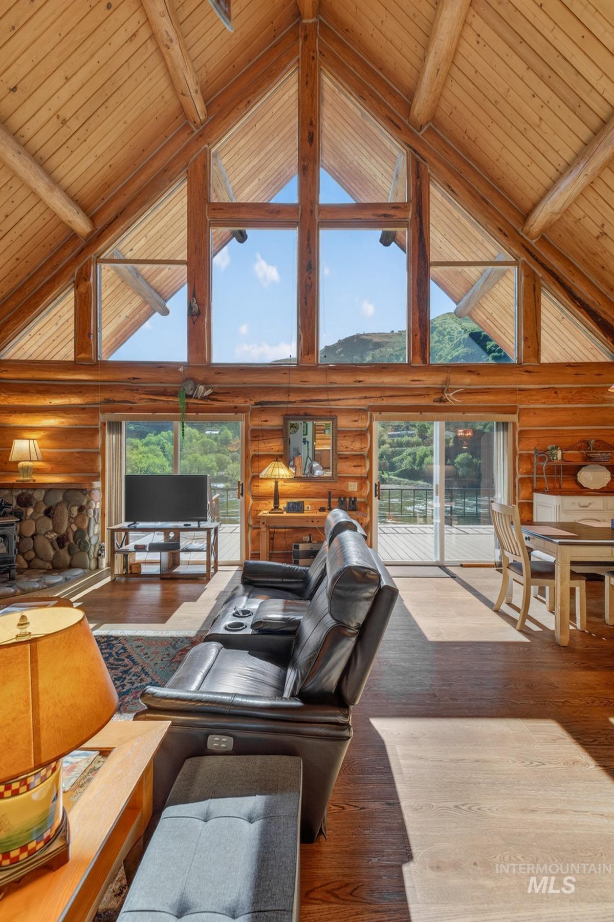 215 Doumecq Road White Bird, ID 83554 - Photo 8 of 50 Vaulted log cabin interior featuring a wall of windows, exposed timber framing, a stone fireplace with a wood stove, and wood-finish flooring