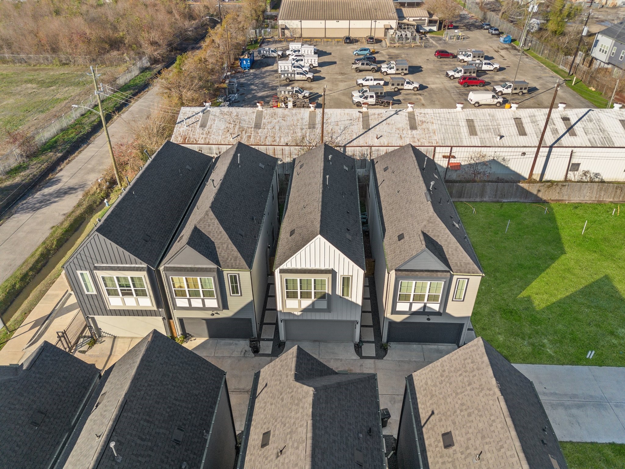 2928 Stevens Street Houston, TX 77026 - Photo 27 of 30 Modern townhomes with dark roofs and large windows are lined up closely, featuring small driveways. The area is adjacent to a commercial parking lot and a green space, providing a mix of urban and residential vibes.