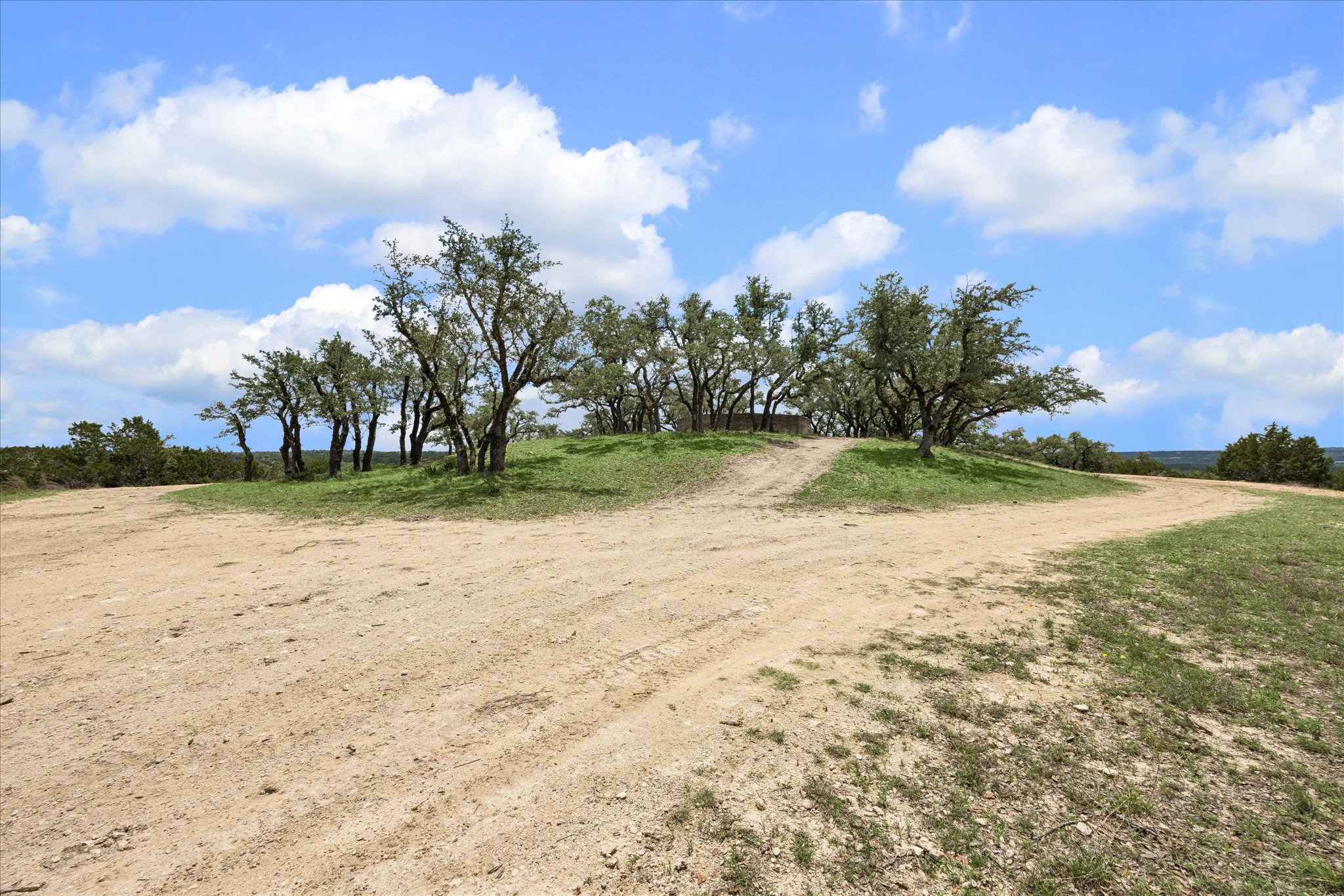 6411 West Fitzhugh Road Dripping Springs, TX 78620 - Photo 22 of 34 Expansive dirt path traversing a natural landscape with scattered mature trees and green ground cover under a blue sky with clouds