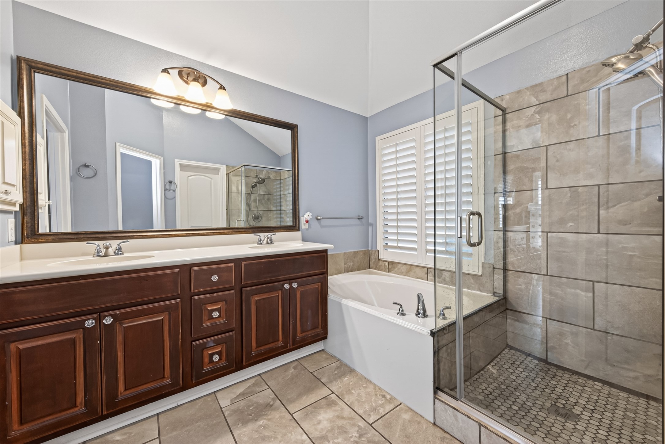 27443 Rebecca Field Lane Spring, TX 77386 - Photo 18 of 31 This bathroom features a spacious layout with a double-sink vanity and dark wood cabinets, a glass-enclosed shower with tiled walls, and a separate bathtub with elegant fixtures. Natural light streams through window shutters, highlighting the neutral tile flooring.