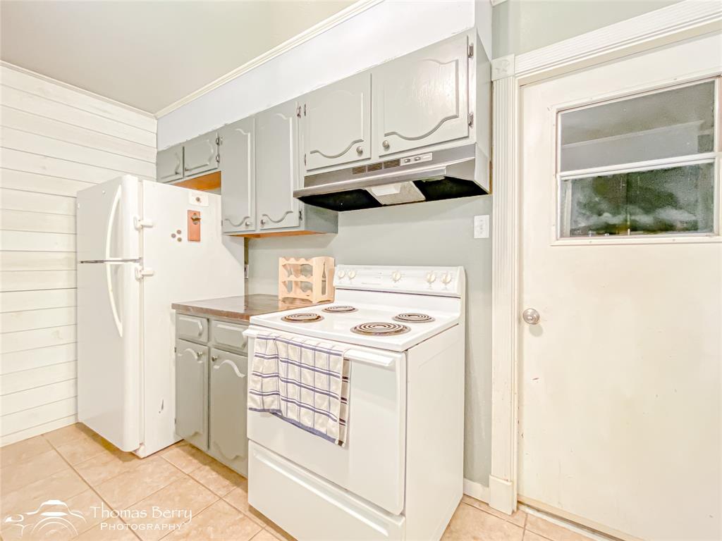 1104 Hickory Street Roscoe, TX 79545 - Photo 11 of 22 Kitchen featuring white appliances, under cabinet range hood, light tile patterned flooring, light countertops, and wooden walls
