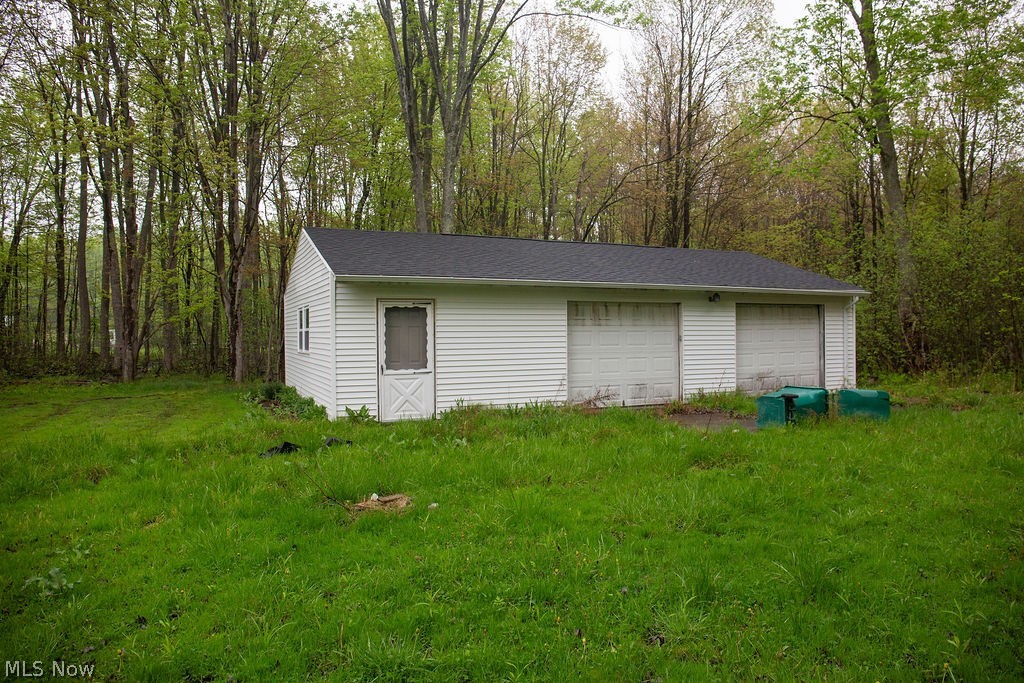 3644 Padanarum Road Geneva, OH 44041 - Photo 18 of 19 Detached garage with white vinyl siding, a dark shingle roof, two garage doors, and a side entry door, surrounded by natural foliage