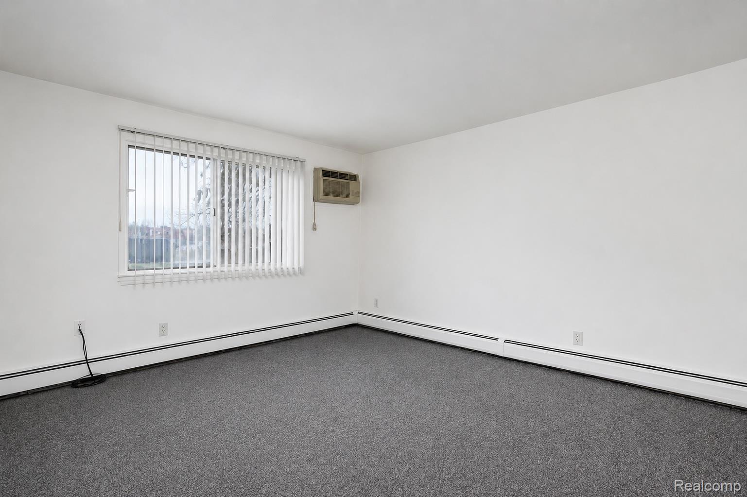 Wall-to-wall carpeting, white painted walls, window with vertical blinds, and a wall-mounted air conditioning unit