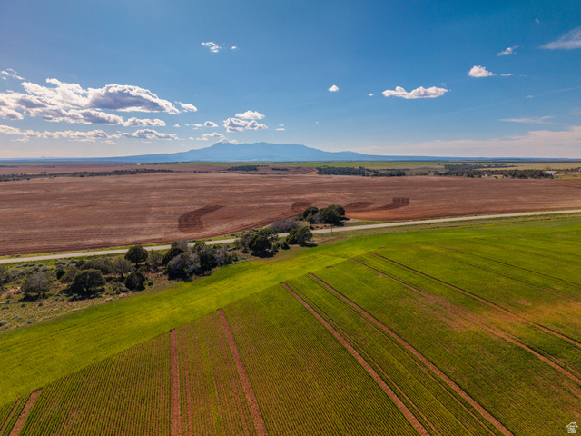 Expansive agricultural land featuring a road, varied green fields, and tilled brown fields, all set against a distant mountain range under a blue sky with scattered clouds