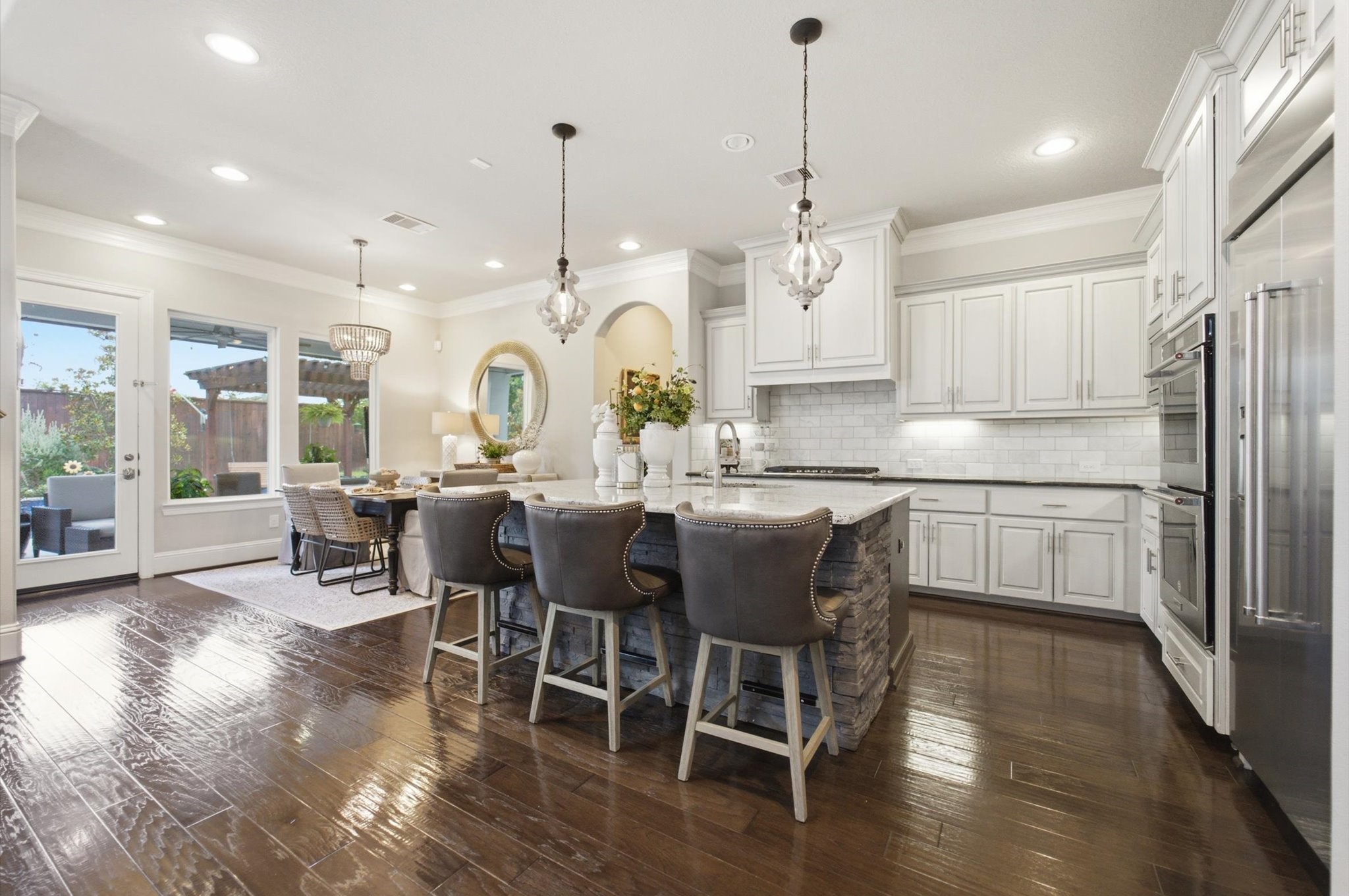 23722 Thortons Park Lane Spring, TX 77389 - Photo 21 of 48 This photo showcases a modern, open-concept kitchen and dining area. It features a large island with stylish barstools, elegant pendant lighting, and stainless steel appliances. The space has hardwood floors, white cabinetry, and a bright, airy atmosphere with large windows leading to an outdoor patio.