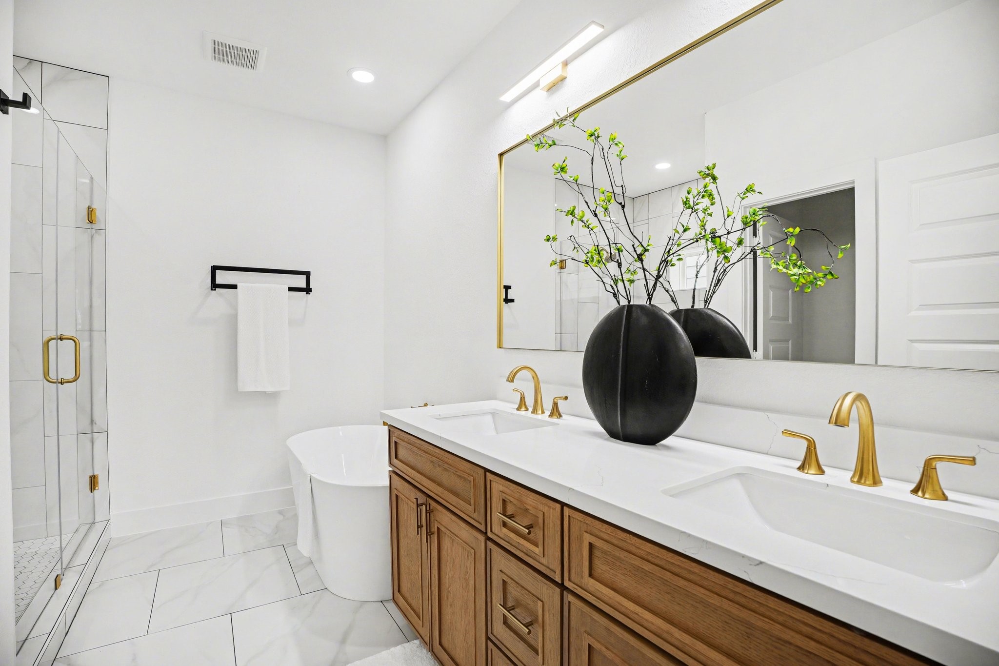 115 Milby Street Houston, TX 77003 - Photo 15 of 28 This modern bathroom features a dual-sink vanity with sleek gold fixtures, a large mirror, and elegant cabinetry. A standalone bathtub and a glass-enclosed shower add luxury, complemented by bright lighting and a clean, minimalist design.