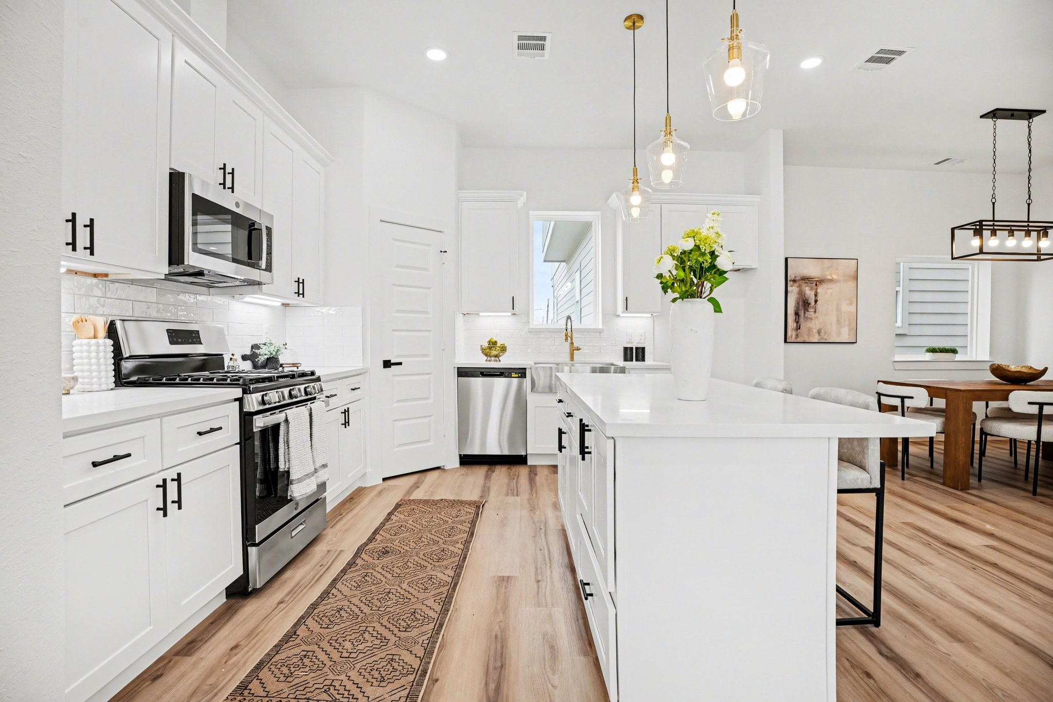 115 Milby Street Houston, TX 77003 - Photo 8 of 28 This kitchen features a modern design with white cabinetry, stainless steel appliances, and a central island with pendant lighting. The open layout seamlessly connects to a dining area with contemporary furnishings and stylish light fixtures, creating an inviting and functional space.