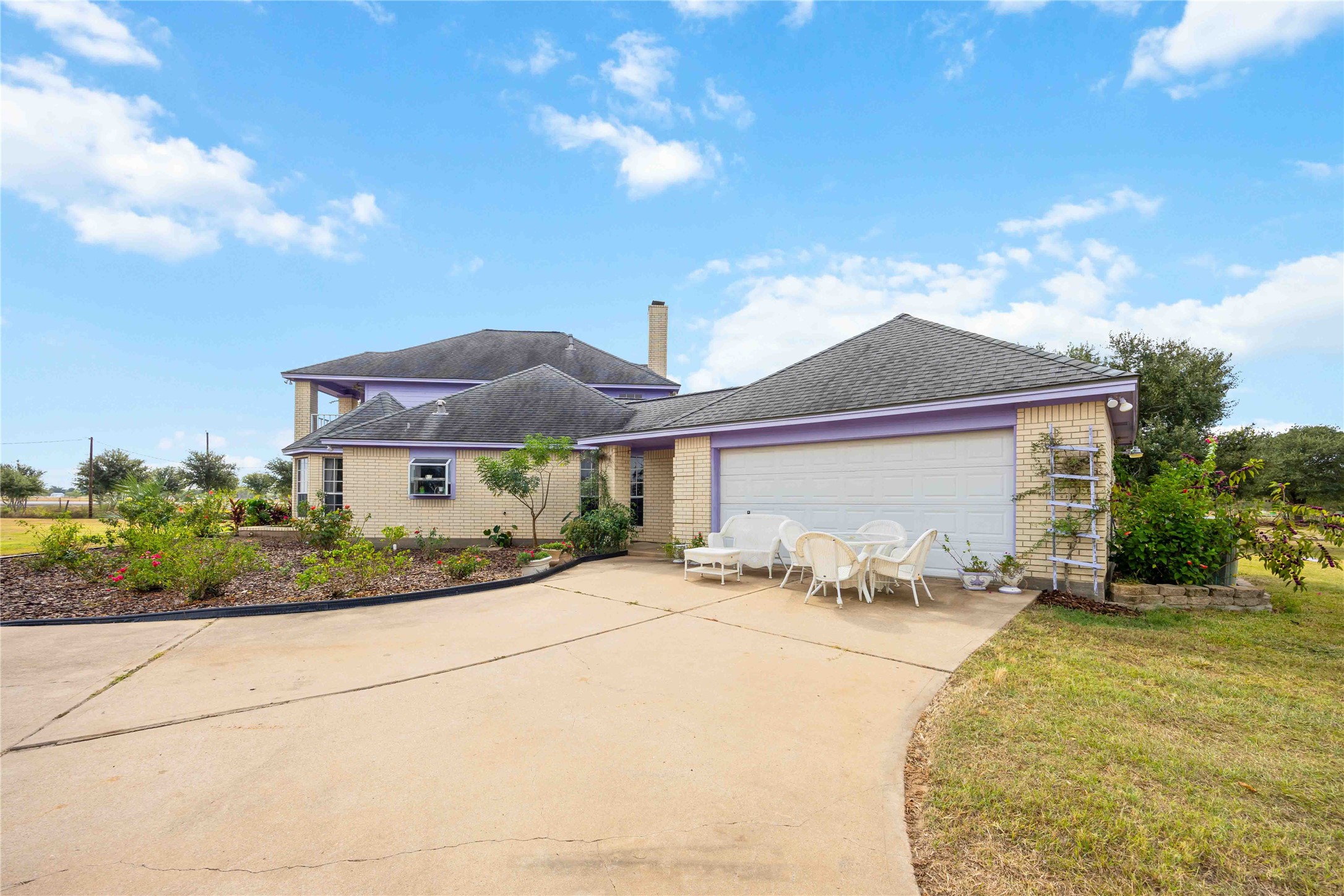 12075 Farm To Market 359 Hempstead, TX 77445 - Photo 14 of 46 This is a charming two-story home with a spacious driveway leading to an attached garage. The exterior features light brick with purple accents, and the front yard is landscaped with plants. A patio area with seating is present, offering a welcoming outdoor space.