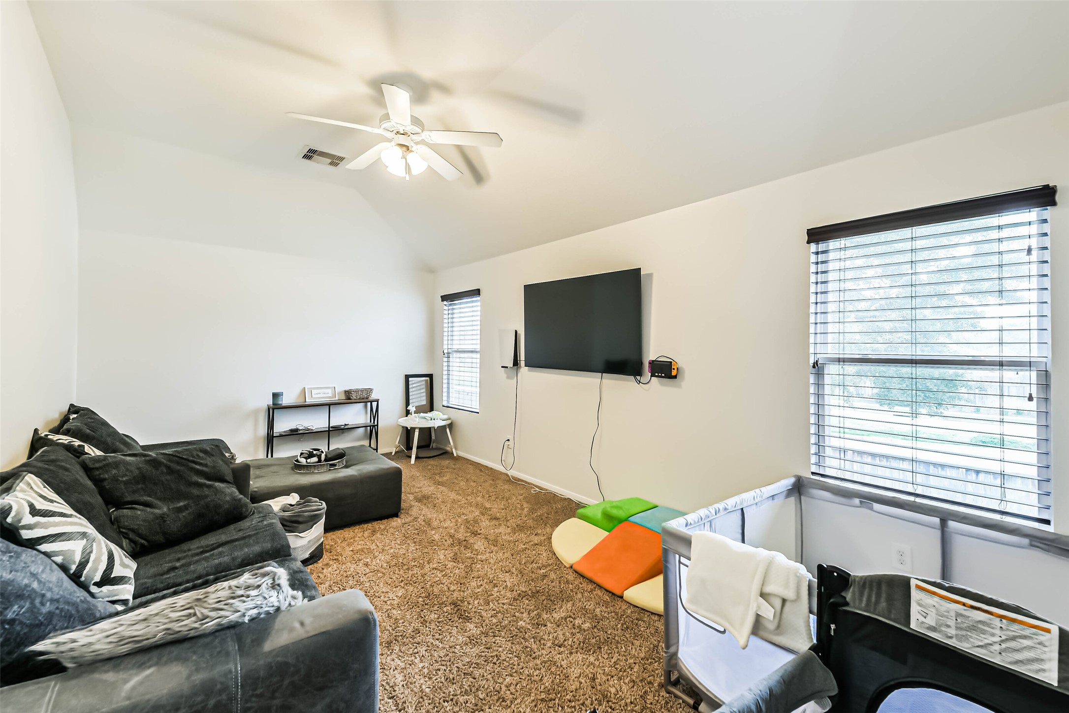 22538 Goss Spring Court Spring, TX 77373 - Photo 21 of 36 This is a cozy living room with neutral walls, featuring a plush carpet, a ceiling fan, and a wall-mounted TV. The space is brightened by two windows with blinds, and it includes a comfortable sofa and a playpen, making it a versatile family area.