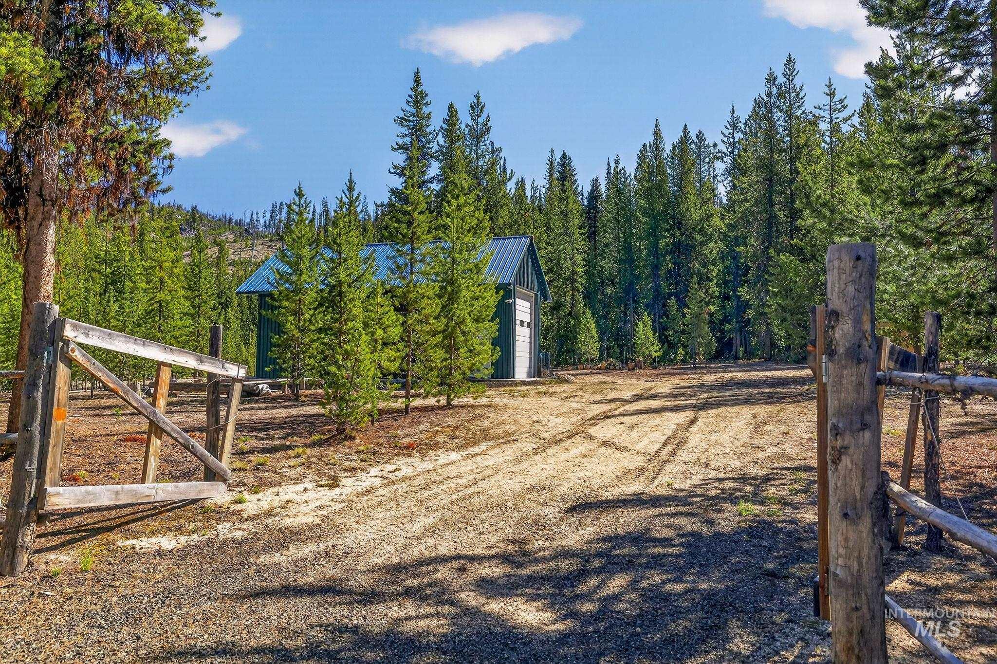 321 Secesh River Road McCall, ID 83638 - Photo 29 of 50 Wood fence entry to a gravel driveway, leading to a storage shed with a blue metal roof, all surrounded by dense evergreen trees