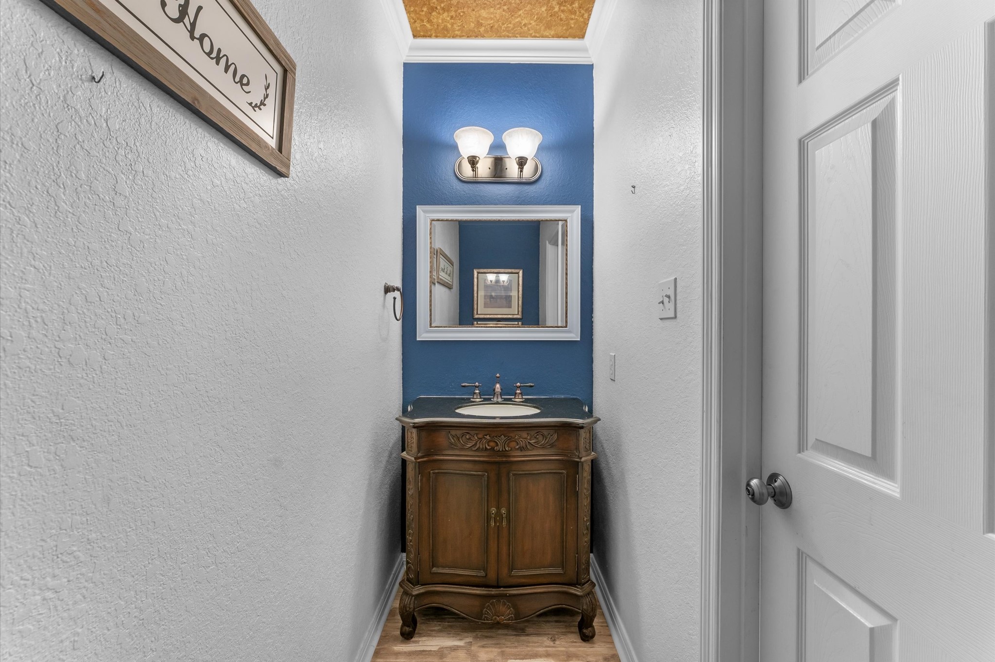 1615 Noble Pointe Drive Spring, TX 77379 - Photo 19 of 50 This photo features a compact powder room with a classic wooden vanity and a blue accent wall. It includes a mirror, dual wall sconce lighting, and a decorative "Home" sign, offering a cozy and welcoming feel.