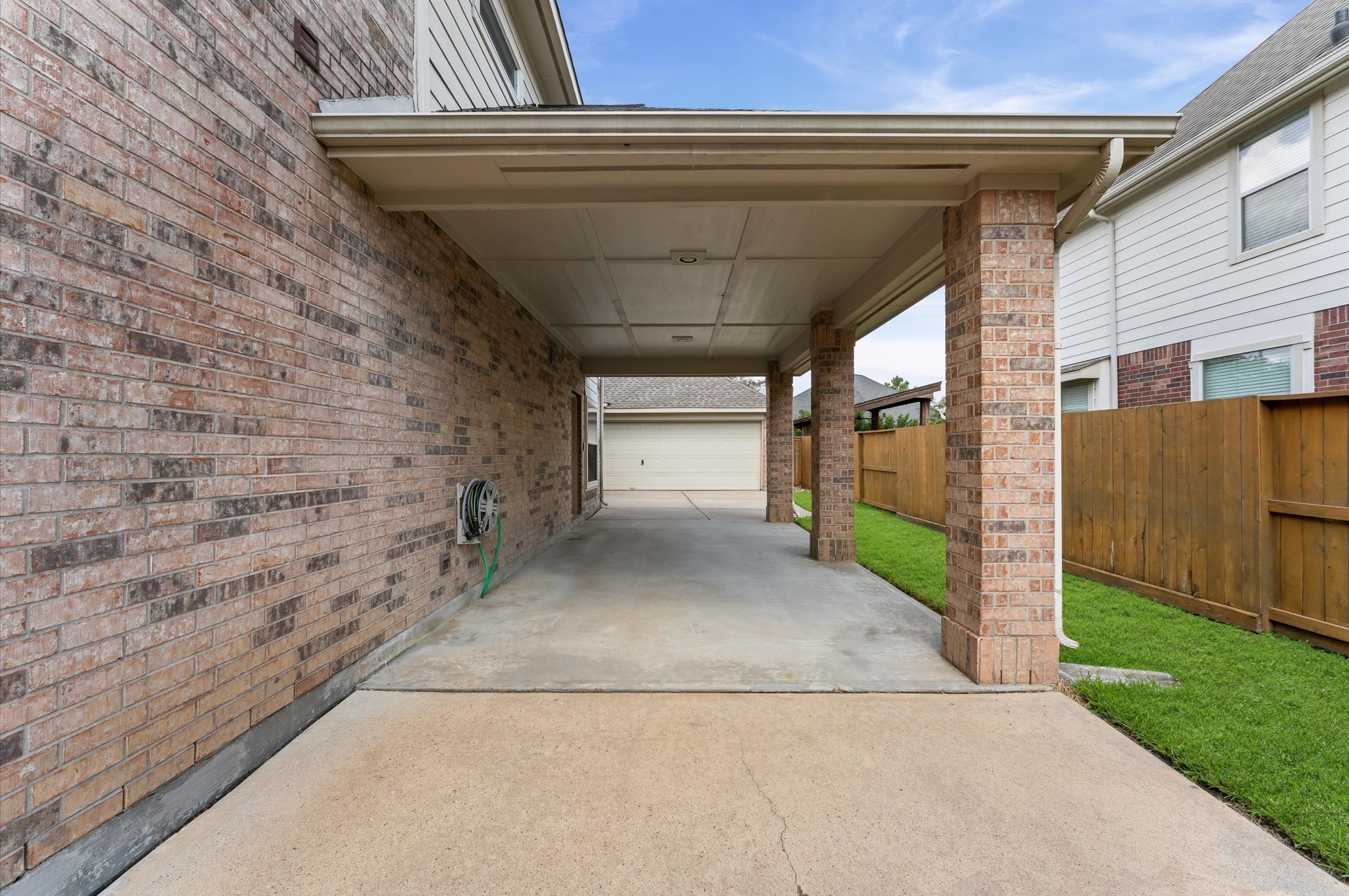 1615 Noble Pointe Drive Spring, TX 77379 - Photo 4 of 50 This photo showcases a spacious covered driveway with brick pillars and a well-maintained concrete surface. It leads to a detached garage, flanked by a brick wall on one side and a wooden fence with a small lawn on the other, providing ample space for parking and outdoor activities.