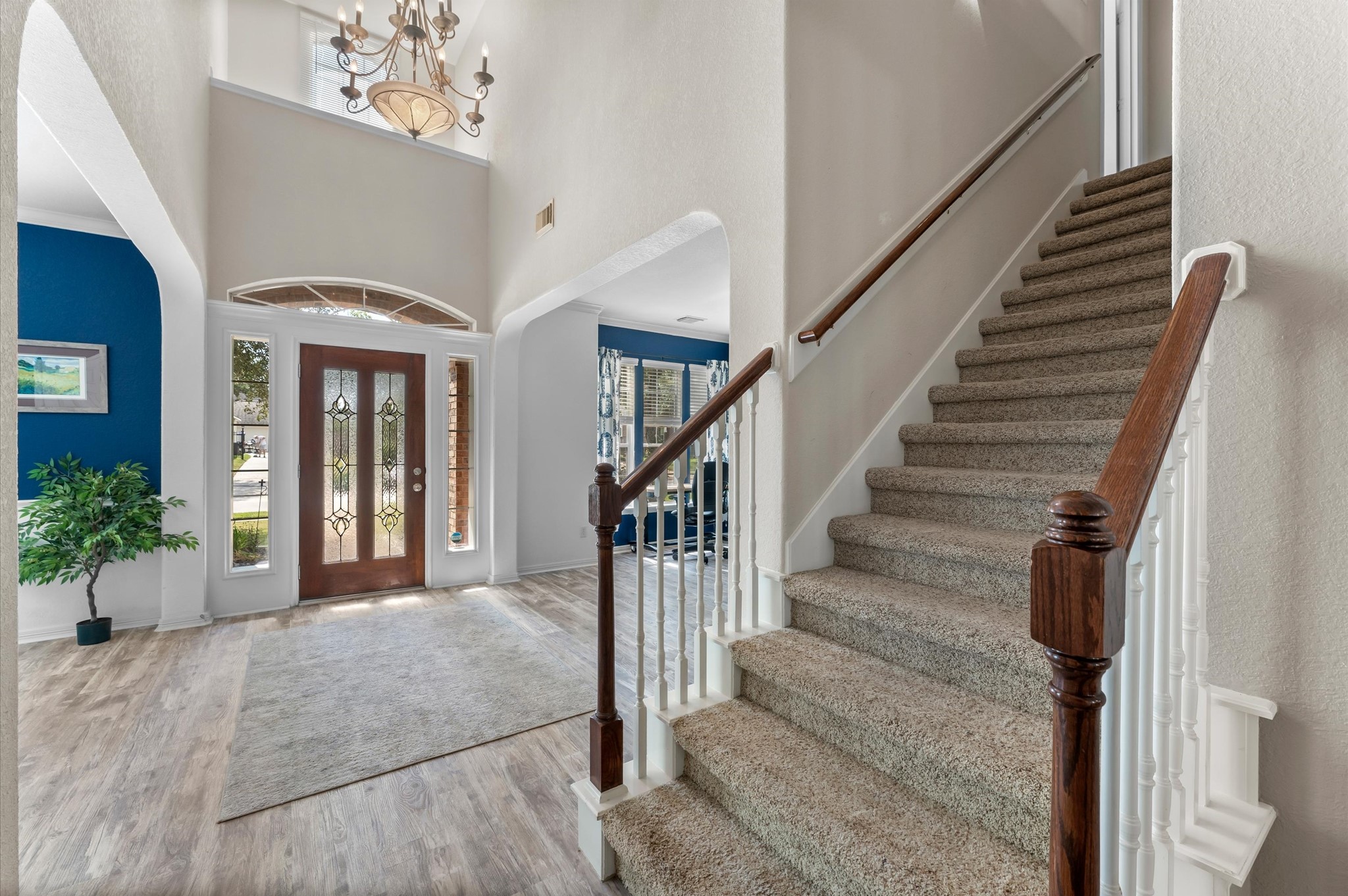 1615 Noble Pointe Drive Spring, TX 77379 - Photo 7 of 50 This welcoming foyer features a high ceiling with a chandelier, a stylish wooden front door with glass panels, and a carpeted staircase with wooden handrails. The entry opens into a bright space with hardwood floors and elegant archways, leading to a room with blue accent walls.