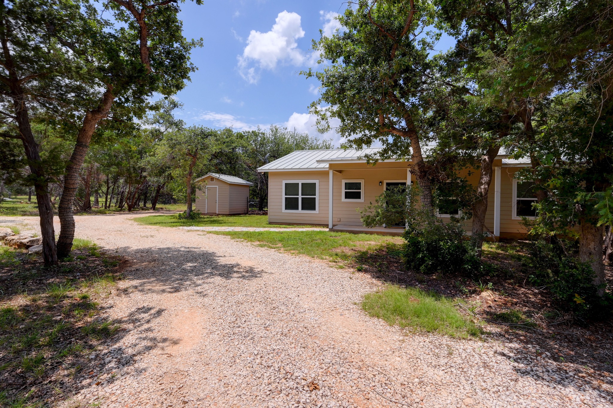 231 North Rainbow Ranch Road Wimberley, TX 78676 - Photo 4 of 40 A quaint residence nestled among mature trees, with a detached shed and a sprawling gravel driveway.