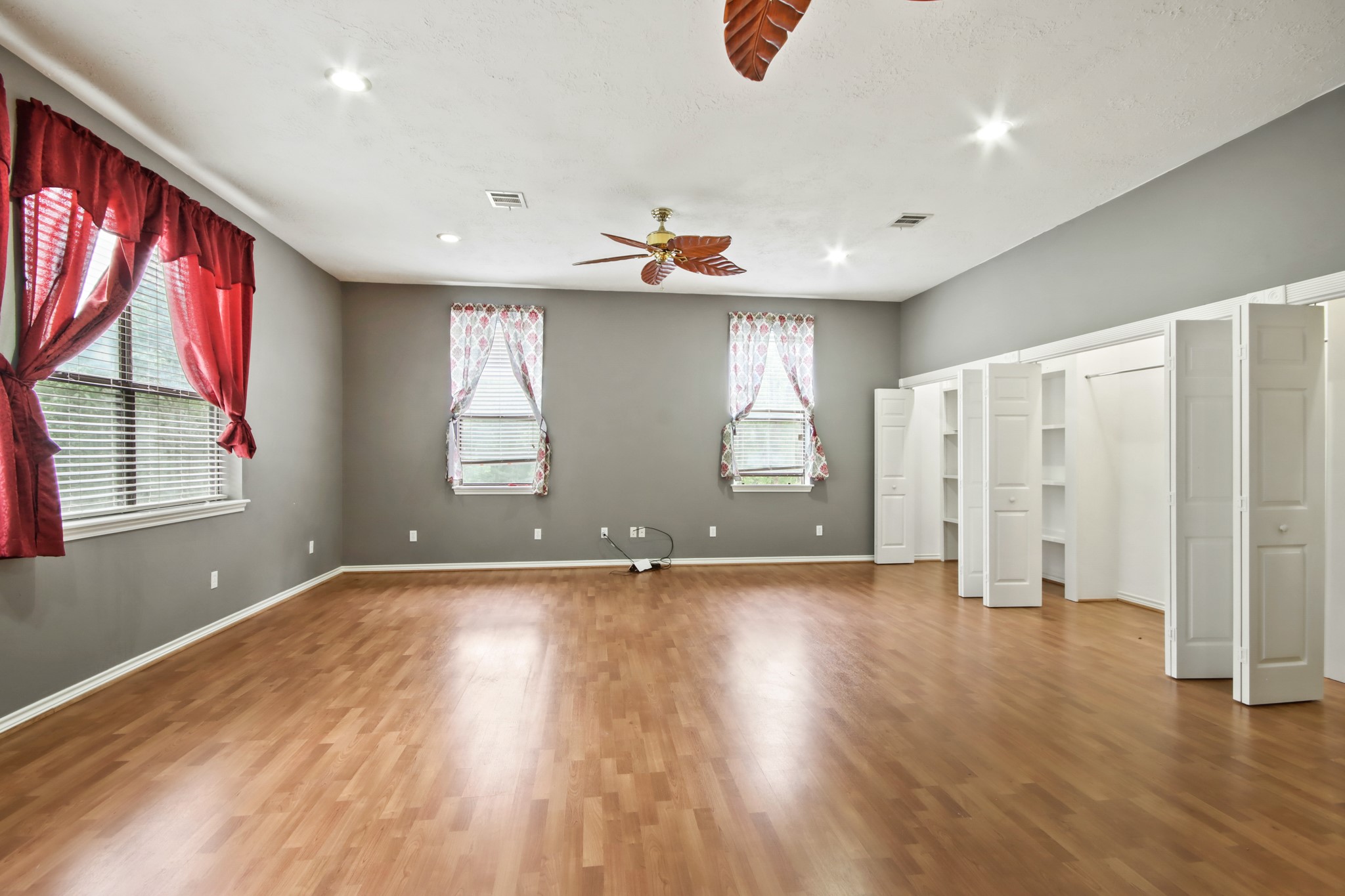 35 Susan Drive Cleveland, TX 77327 - Photo 17 of 33 This spacious room features warm wood flooring, neutral gray walls, and ample natural light from multiple windows with red and patterned curtains. It includes ceiling fans with leaf-shaped blades and built-in shelving for storage.