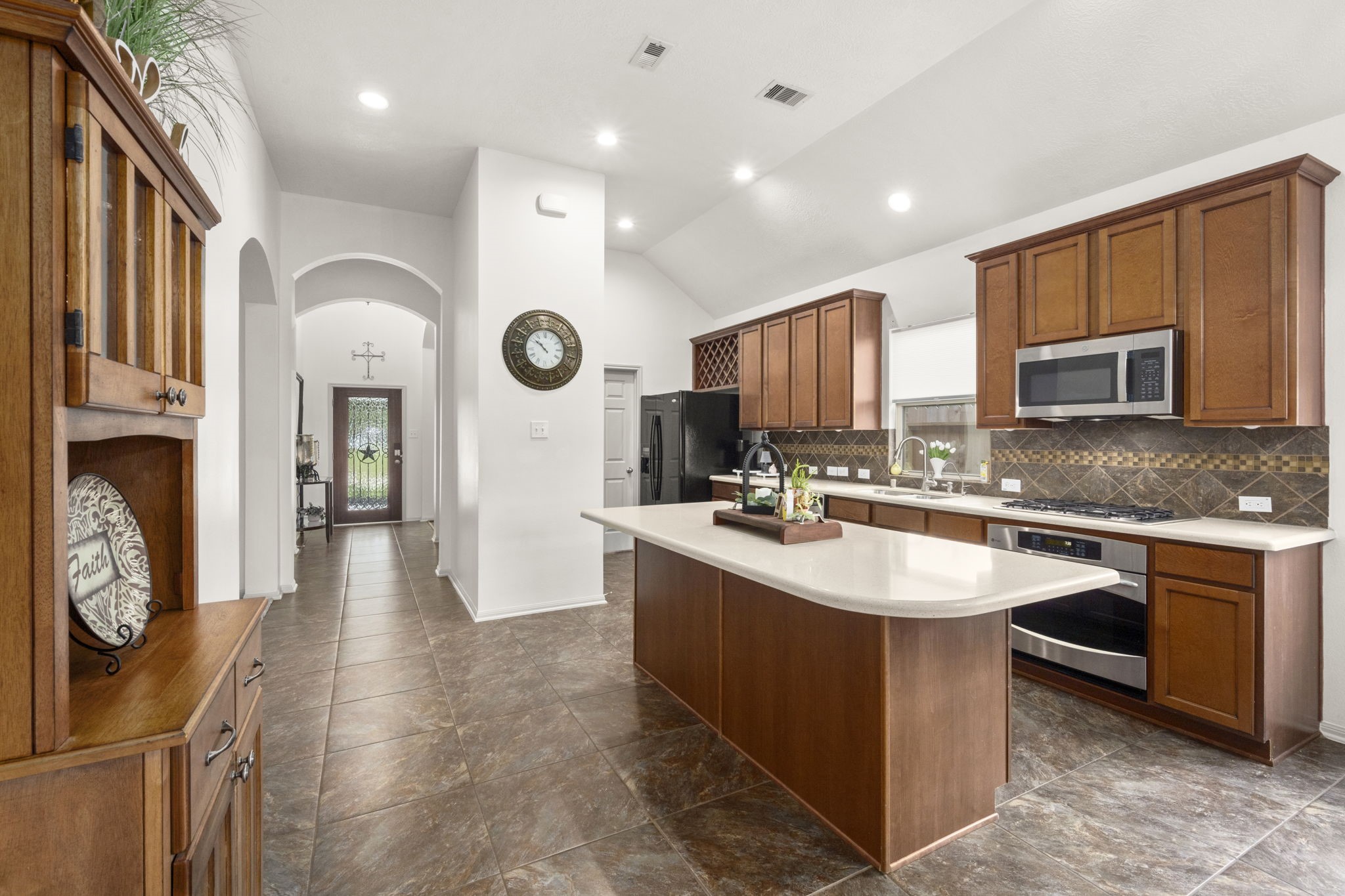 27526 Gatlin Lane Spring, TX 77386 - Photo 8 of 31 The inviting kitchen features elegant wood cabinetry, a large island with a sink, stainless steel appliances, and stylish tile flooring. The open layout flows into a bright hallway with arched doorways and modern lighting.
