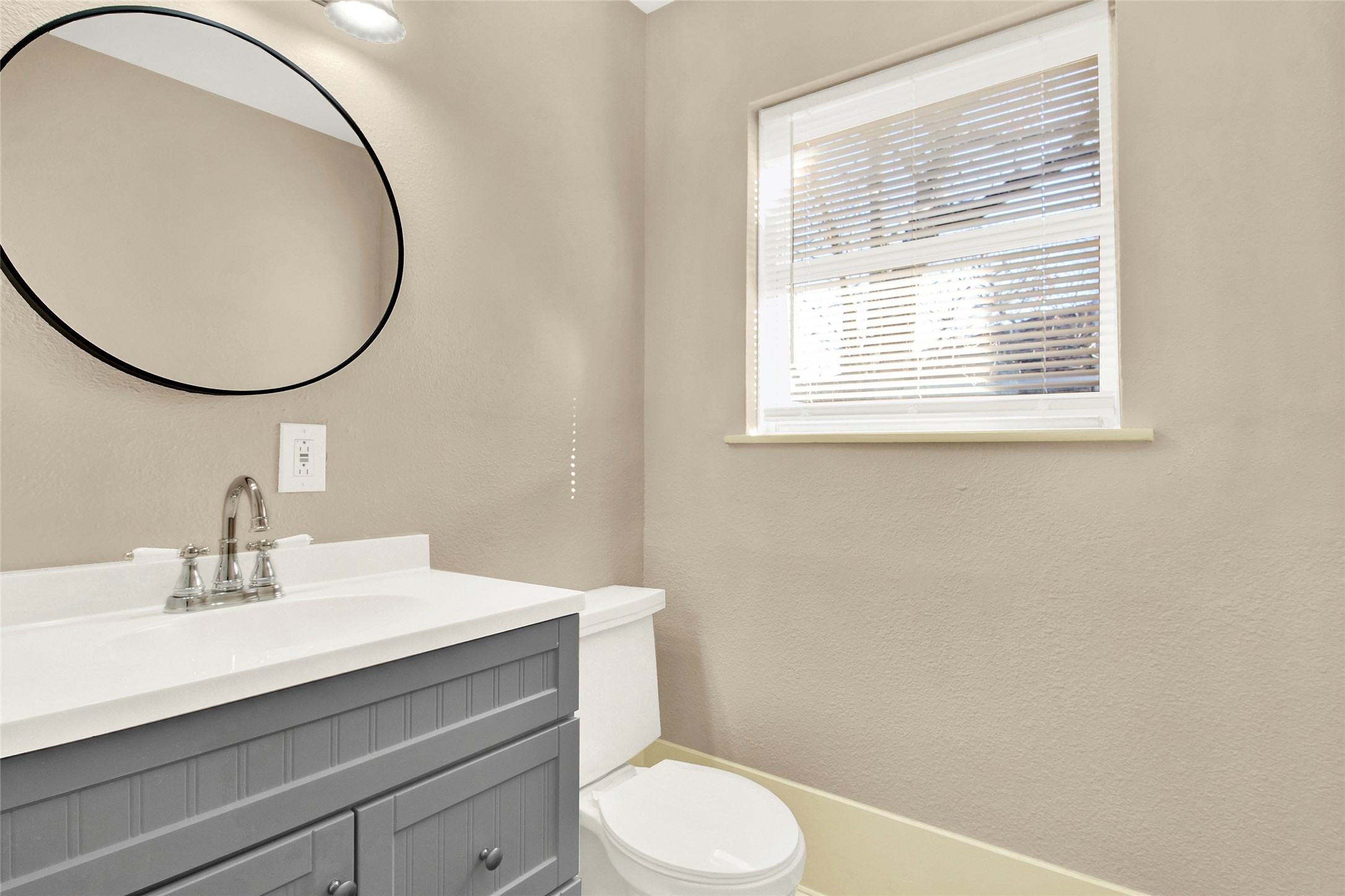 506 Edgebrook Drive Houston, TX 77034 - Photo 15 of 18 This photo shows a modern bathroom with neutral-colored walls, a sleek gray vanity with a white countertop and a stylish round mirror. There's a window with blinds allowing natural light to brighten the space.