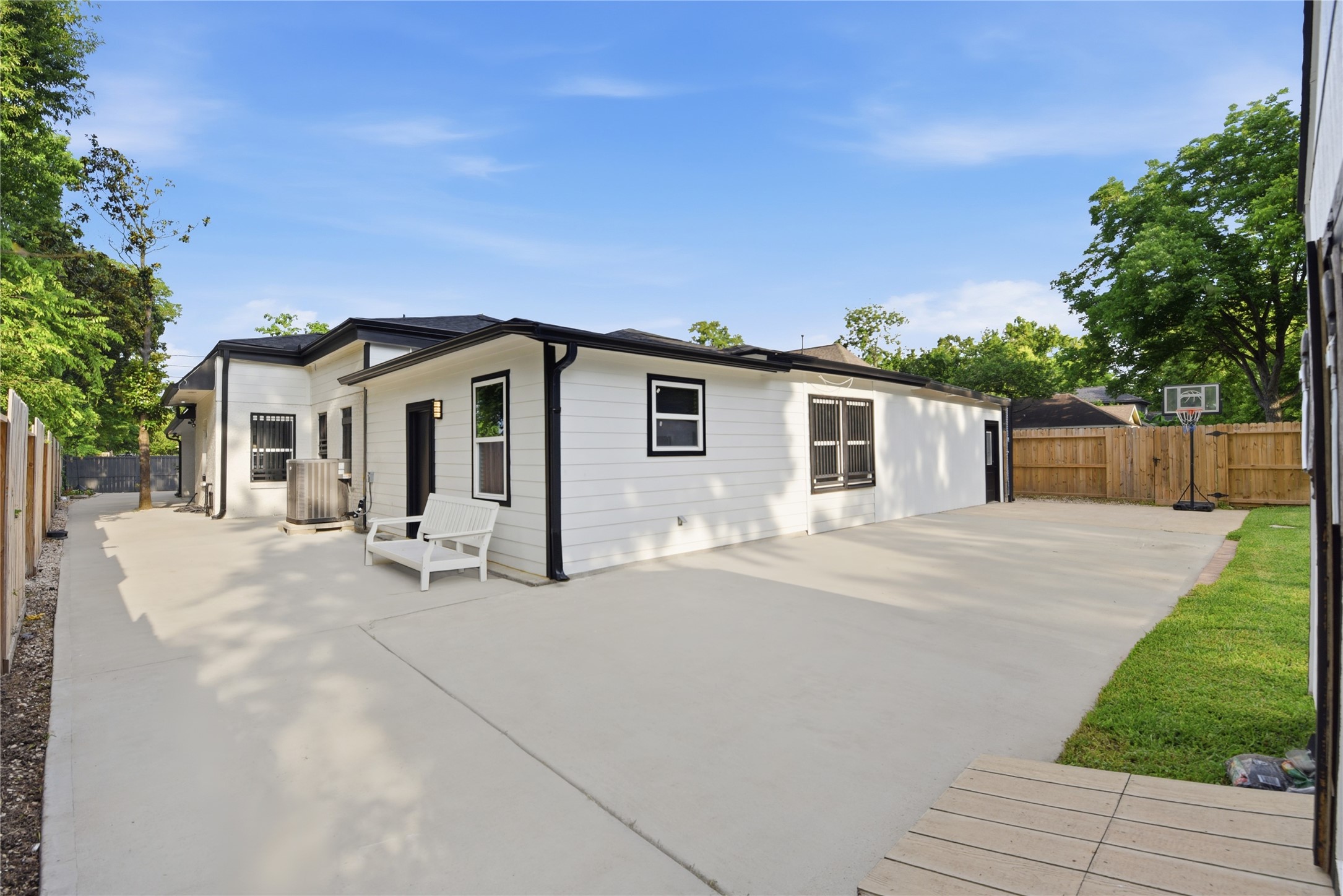 9403 Spaulding Street Houston, TX 77016 - Photo 2 of 50 This photo showcases a modern backyard with a spacious concrete patio, white siding, and black trim. It includes a small grassy area, basketball hoop, and privacy fencing, perfect for outdoor activities and relaxation.