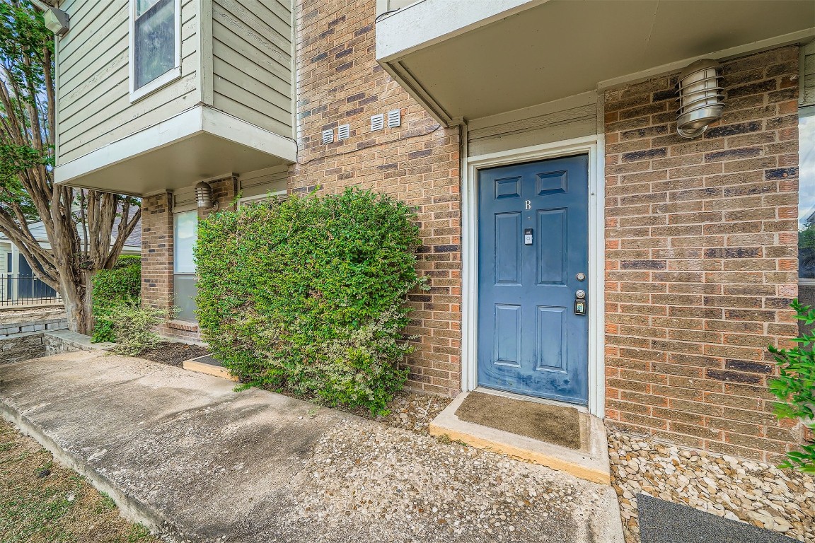 2018 West Rundberg Lane, Unit 2B Austin, TX 78758 - Photo 5 of 31 This photo shows a townhouse entrance with a blue door, surrounded by brick and siding. There's a small, neat shrub adjacent to a concrete walkway, with a modern outdoor light fixture above.