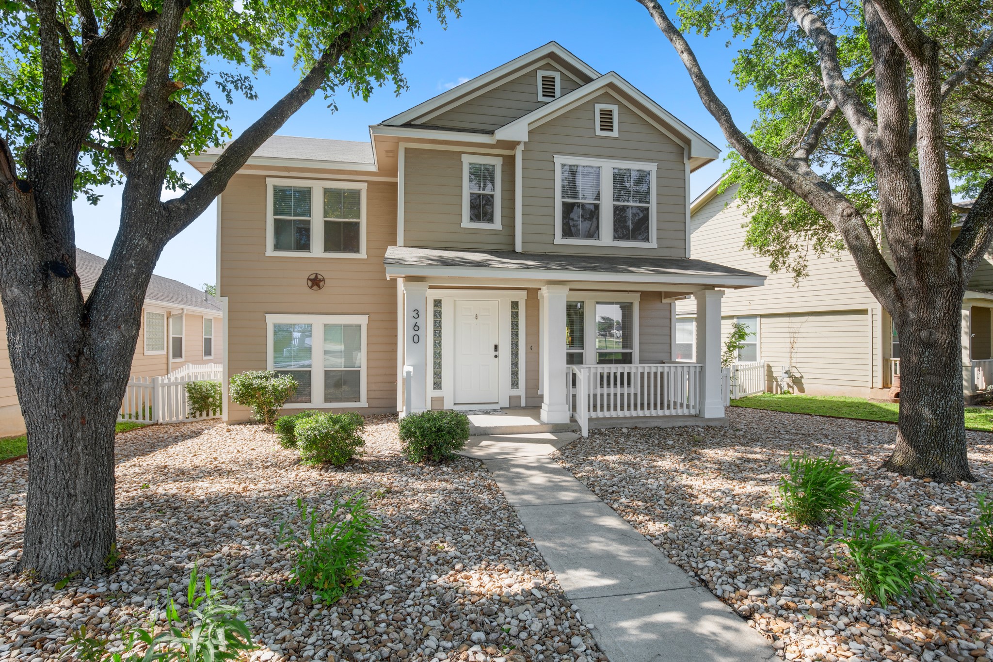 Two-story residence featuring a covered front porch with white railings, a stone-accented front yard, and mature trees framing the property