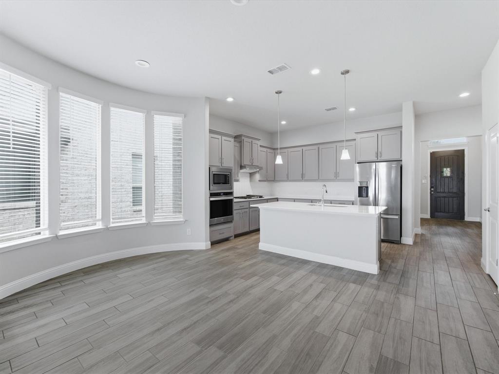 1121 Knightsbridge Road Argyle, TX 76226 - Photo 6 of 39 Open-concept living space featuring wood-look tile flooring, a kitchen island with pendant lighting, a suite of stainless steel appliances, contemporary gray cabinetry, and a curved wall of multiple windows