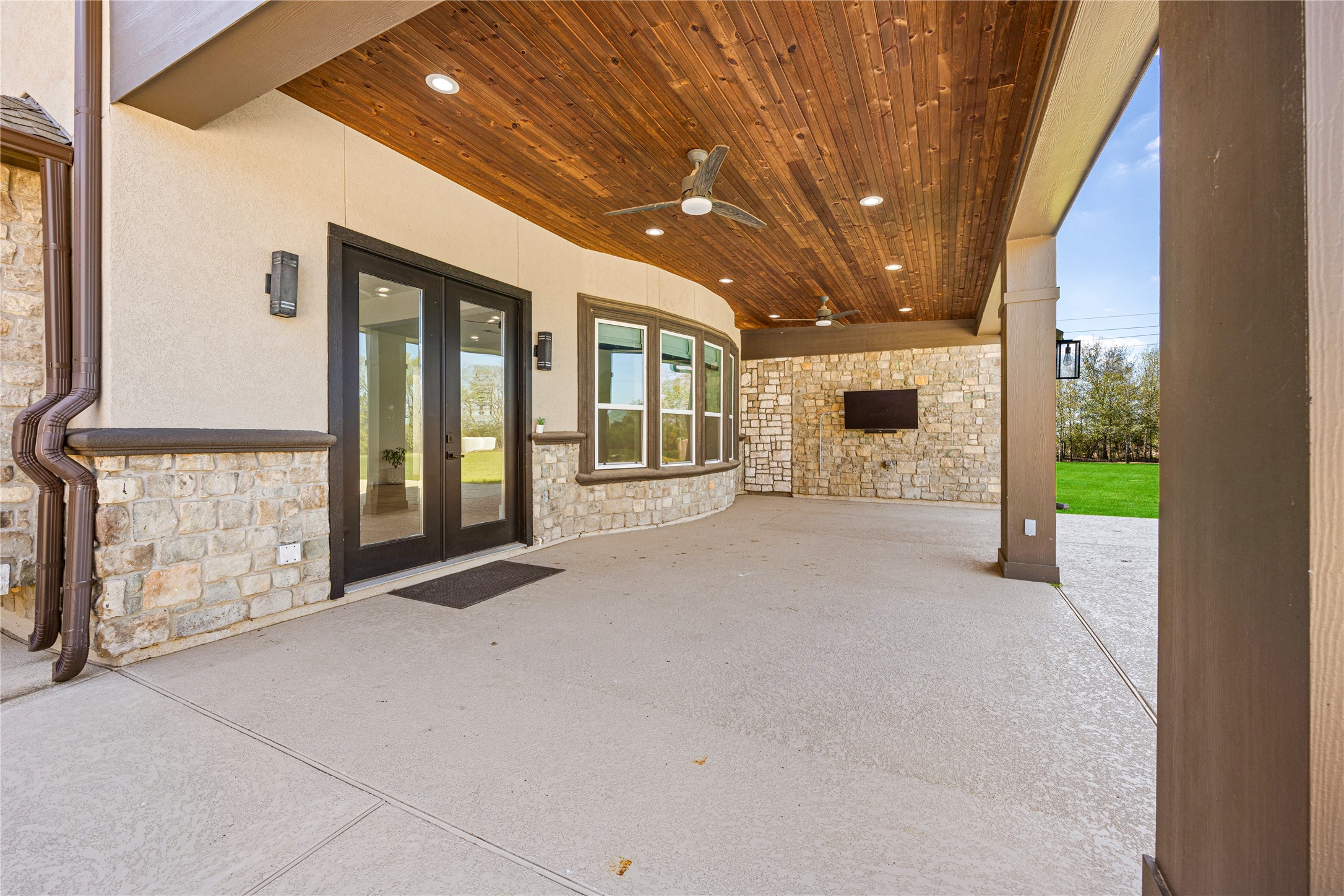 15015 House Road Cypress, TX 77433 - Photo 9 of 50 This photo showcases a spacious covered patio with stylish stone walls, a wood-paneled ceiling, ceiling fans, and a mounted TV, offering a perfect outdoor entertainment space overlooking a lush lawn.