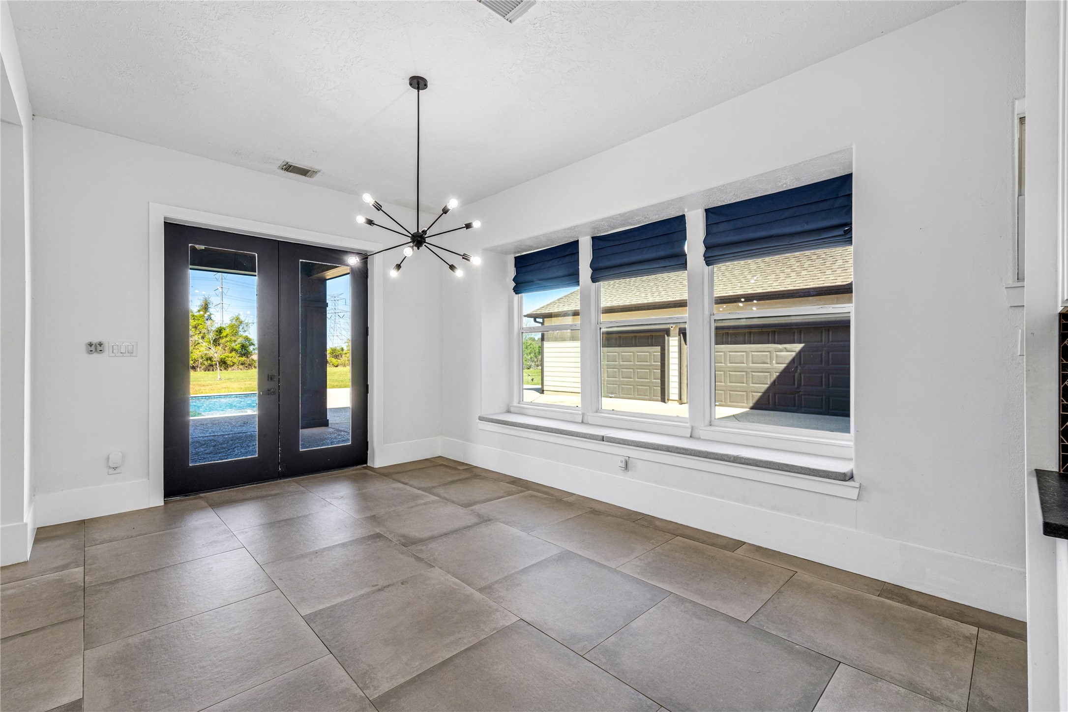 15015 House Road Cypress, TX 77433 - Photo 17 of 50 This spacious room features large tile flooring, a modern chandelier, and wide windows with blue shades. Double doors open to a view of the pool area, providing abundant natural light and a seamless connection to the outdoors.