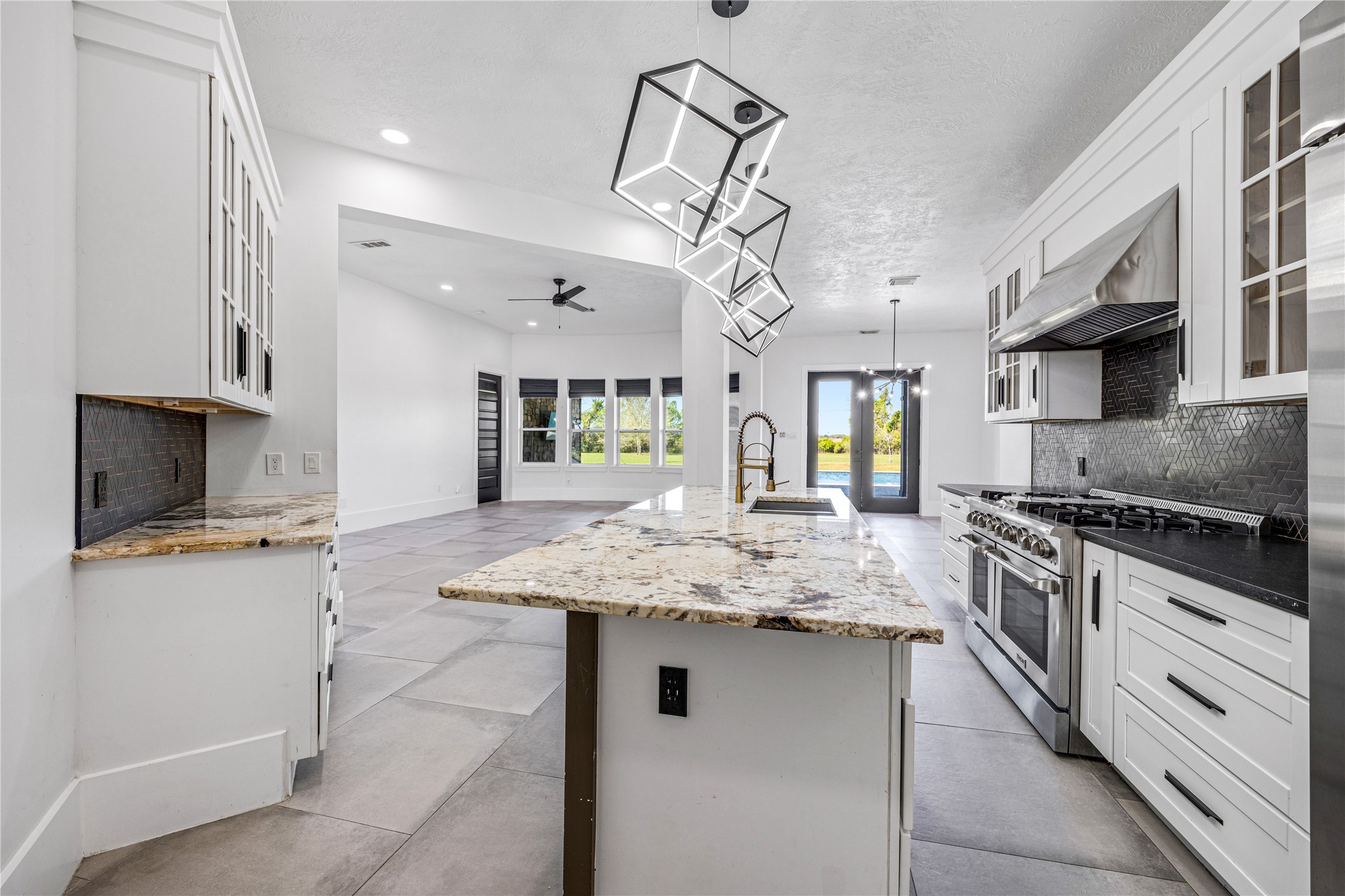 15015 House Road Cypress, TX 77433 - Photo 21 of 50 This photo showcases a modern kitchen with sleek white cabinetry, granite countertops, and a large island. It features stainless steel appliances, including a gas range and hood. The open layout extends into a bright living area with large windows, offering views of the outdoors. Contemporary pendant lights and tile flooring complete the elegant design.