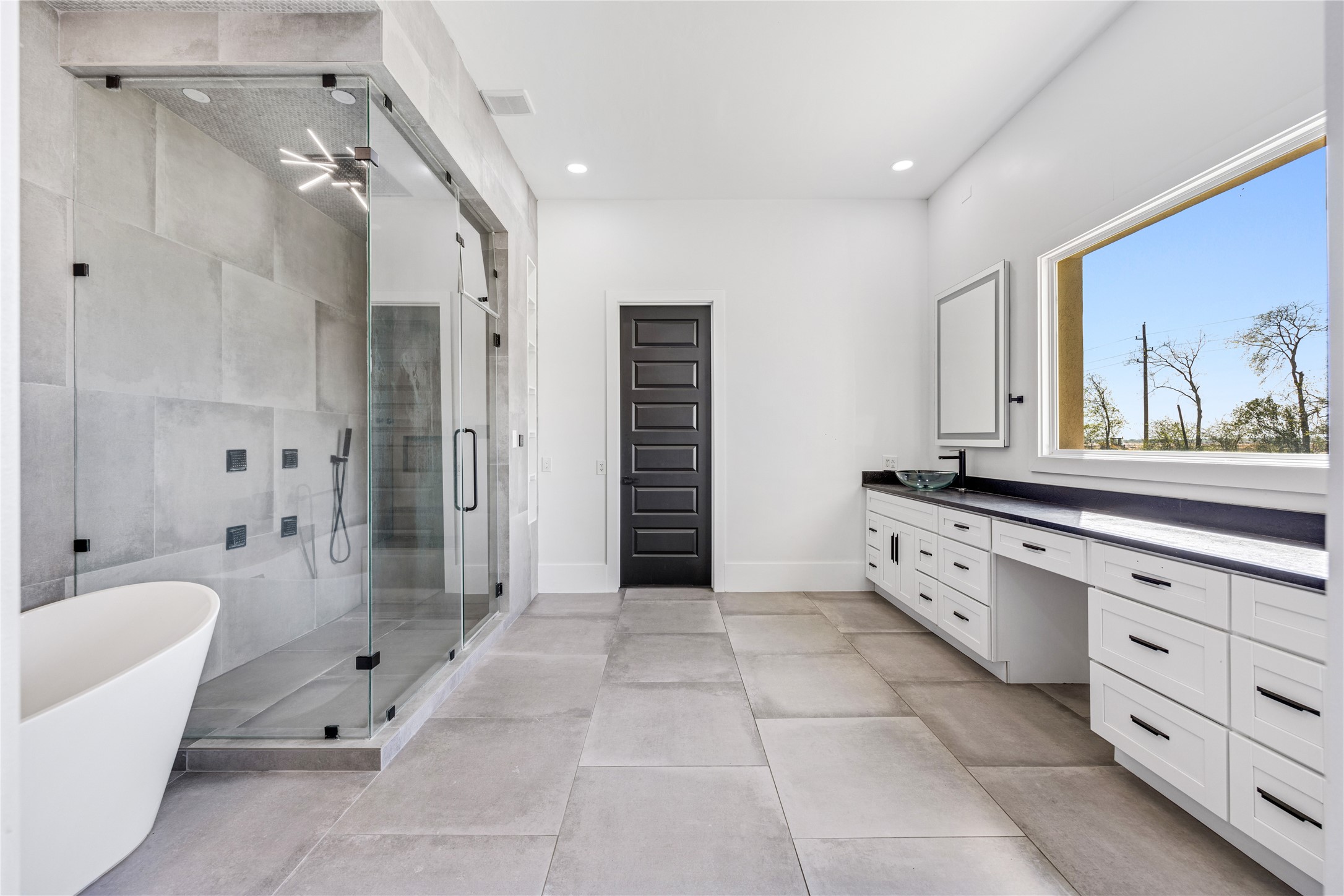 15015 House Road Cypress, TX 77433 - Photo 28 of 50 This bathroom features a spacious layout with a large glass-enclosed shower, a freestanding bathtub, and modern fixtures. It includes a lengthy vanity with ample storage and a window offering natural light. The neutral tile flooring and sleek design create a contemporary feel.