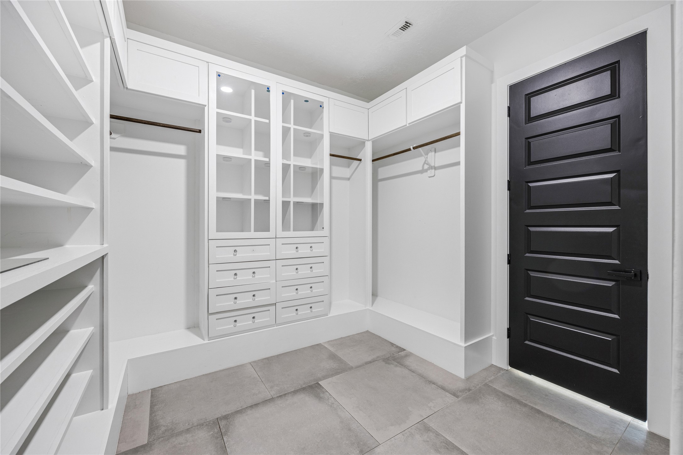 15015 House Road Cypress, TX 77433 - Photo 31 of 50 This spacious walk-in closet features white built-in shelving and cabinets with glass doors, ample hanging space, and a sleek black door, all set on large, light-colored tile flooring. Ideal for organized storage and elegant design.