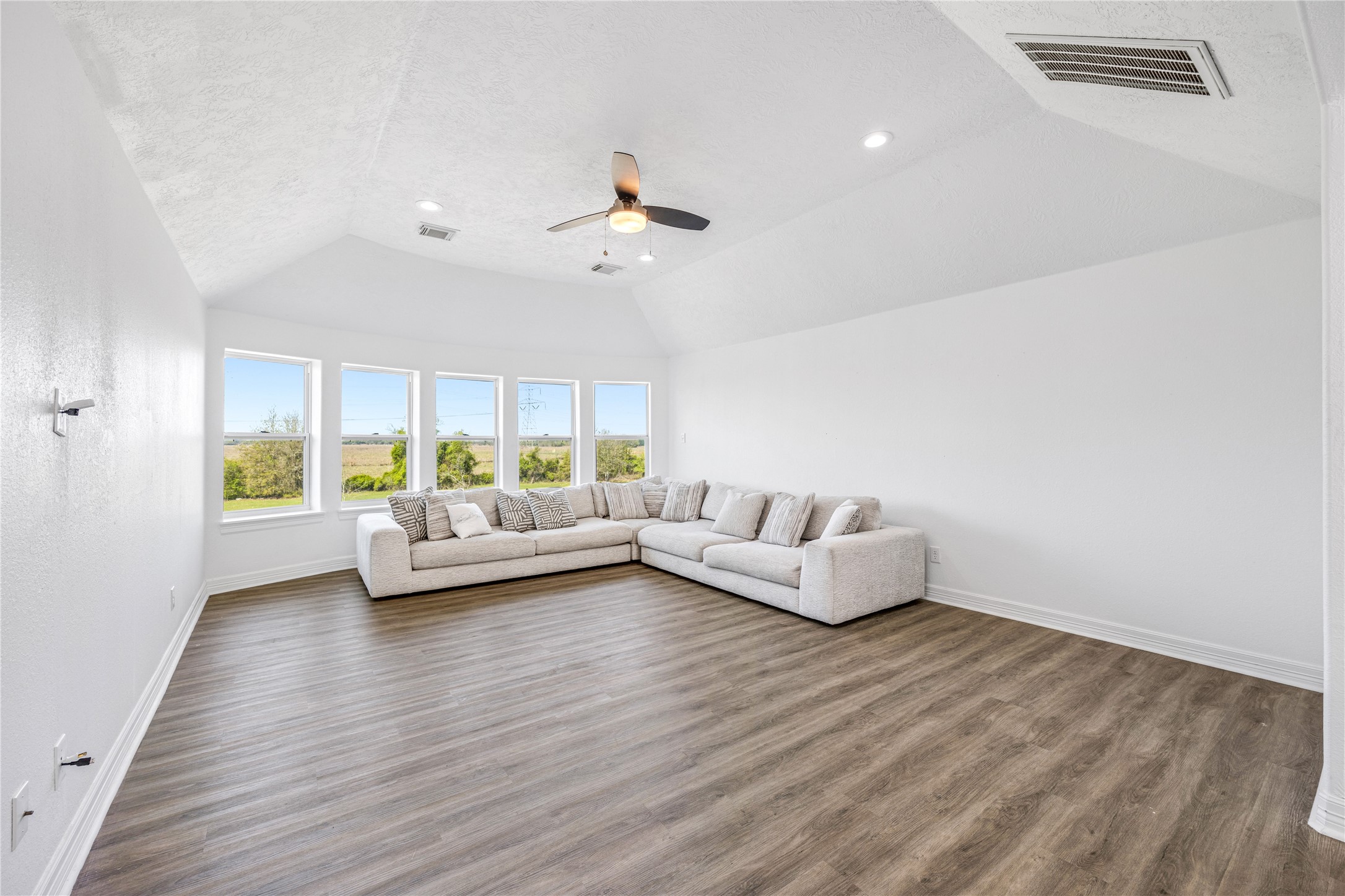 15015 House Road Cypress, TX 77433 - Photo 32 of 50 This spacious living room features a vaulted ceiling, large windows with scenic views, and wood-look flooring. A cozy sectional sofa adds comfort, while a ceiling fan provides a modern touch.