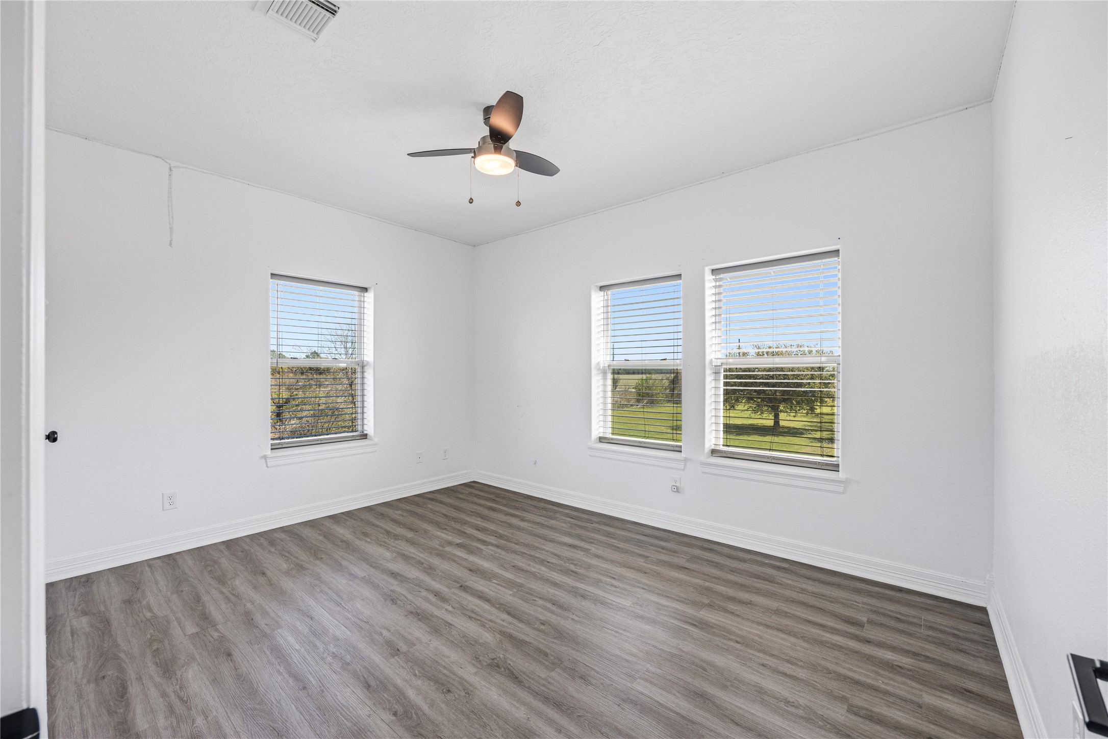 15015 House Road Cypress, TX 77433 - Photo 42 of 50 This room features fresh white walls, a modern ceiling fan, and large windows with views of a lush green landscape, enhanced by sleek wood-look flooring and ample natural light.